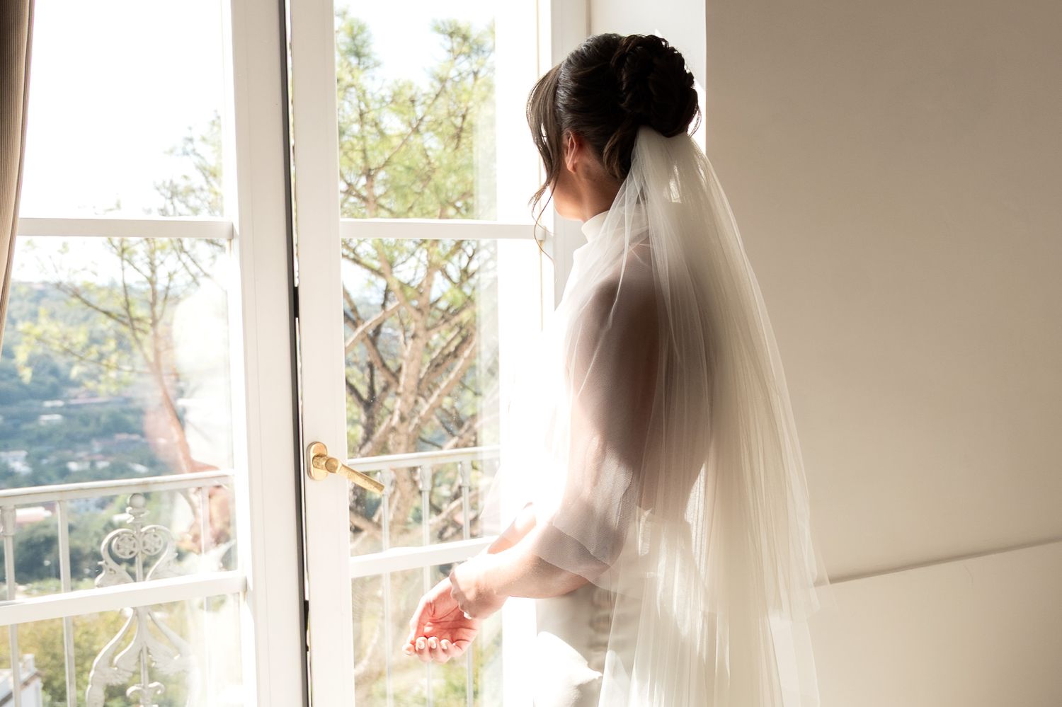 A bride wearing a long veil looking out of a window, she appears deep in thought and adjusts the bracelet on her wrist.