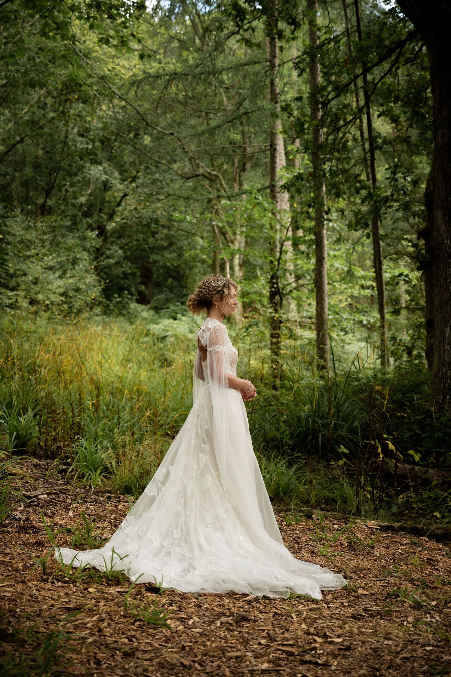 A lady wears a white elegant detailed dress and stands in side profile, she is alone in a forest with her hands clasped in front of her.