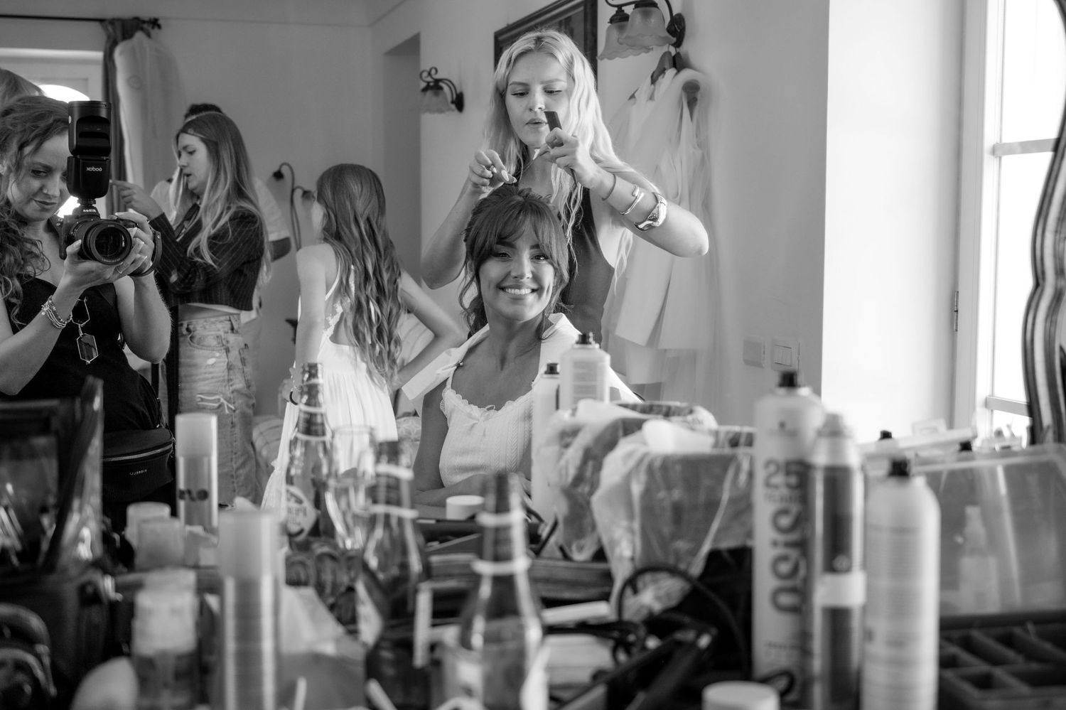 Woman smiles at a photographer in the reflection of a mirror whilst she has her hair styled, she is surrounded by make up and hair products.