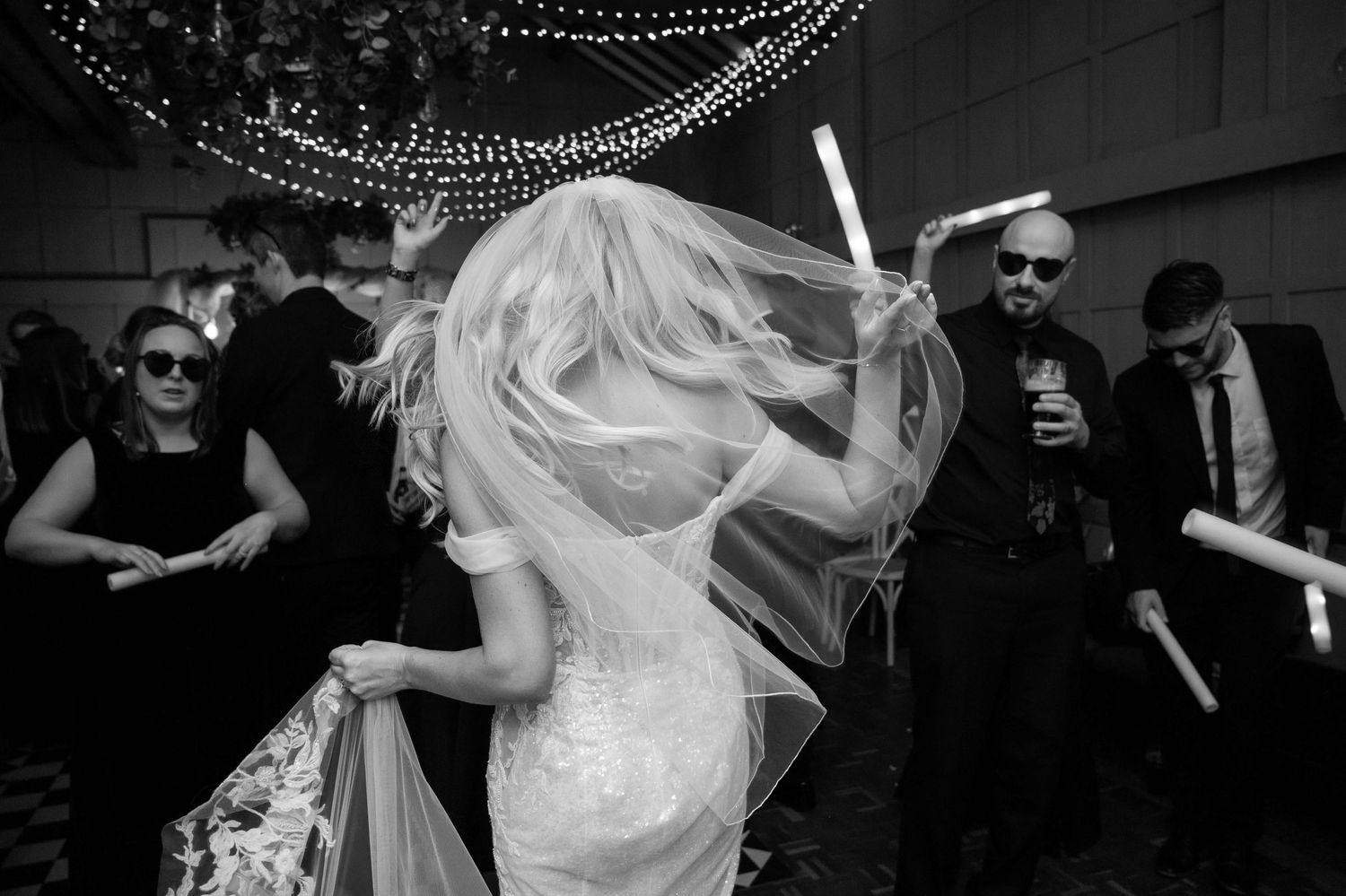 A bride waving a glow stick and dancing energetically with friends who are wearing heart sunglasses. Her hair and veil swish as she moves.