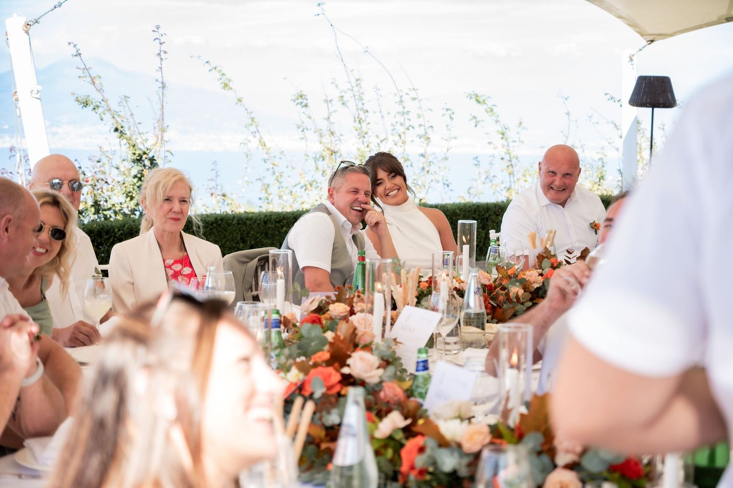 A couple sit closely together at a floral and candle covered table. Friends join them smiling and laughing whilst listening to a speech.