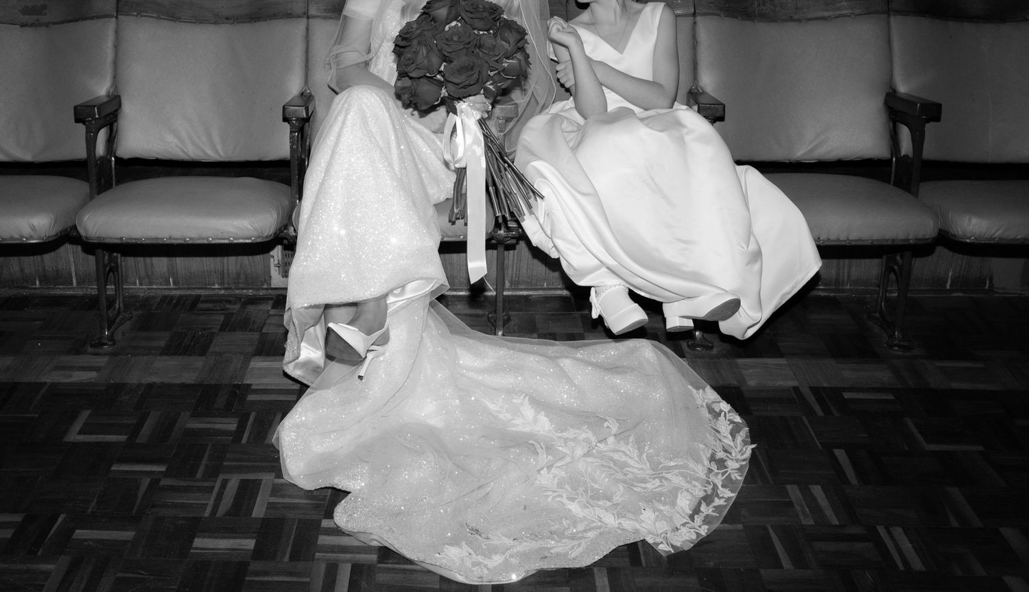A young girl sits next to her mother on cinema style seating, her ribbon shoes swing as she is not tall enough to reach the floor.