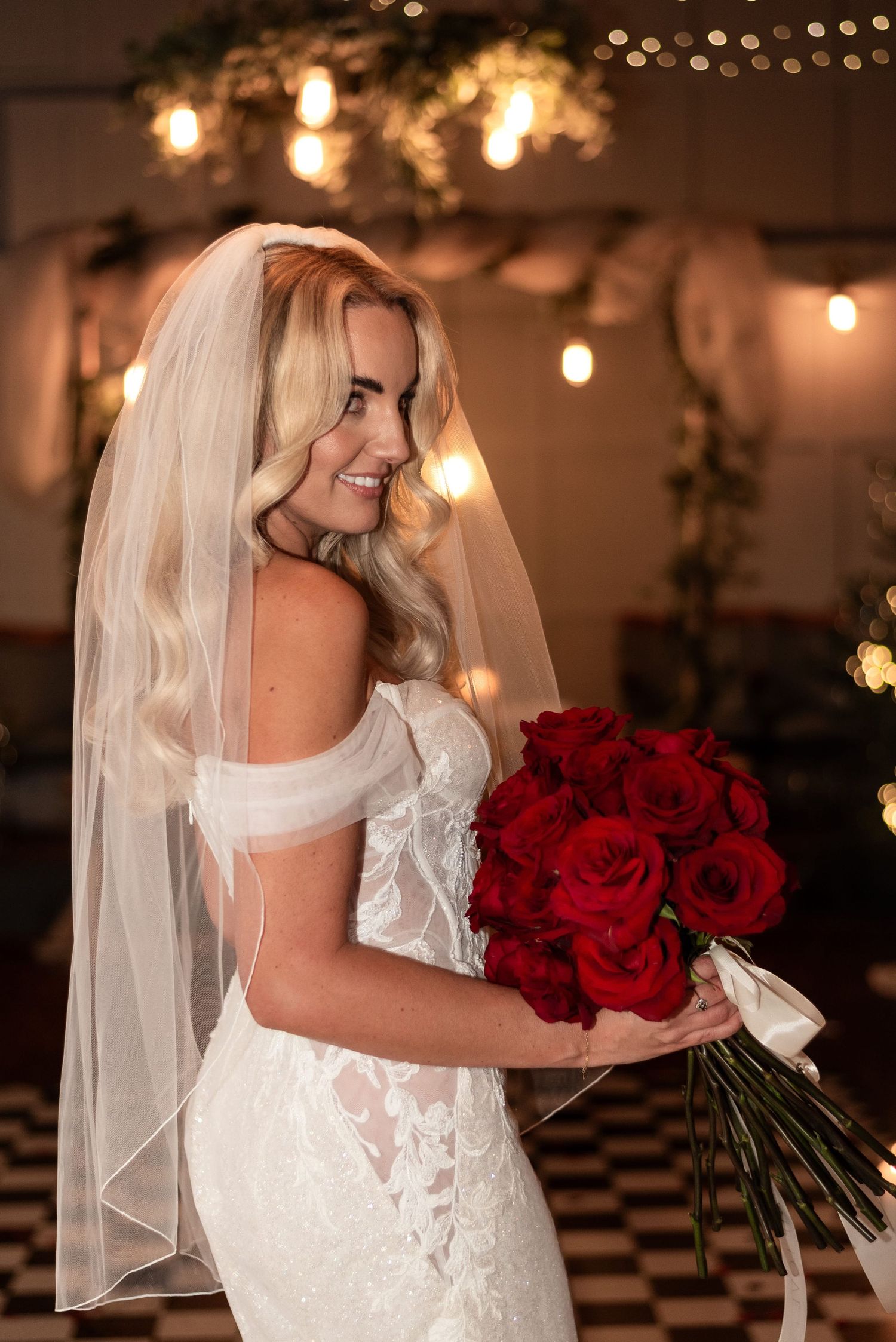Side profile bride in a delicate low shoulder lace and sequin gown, radiant with a wide smile and clutching a  deep red rose bouquet.