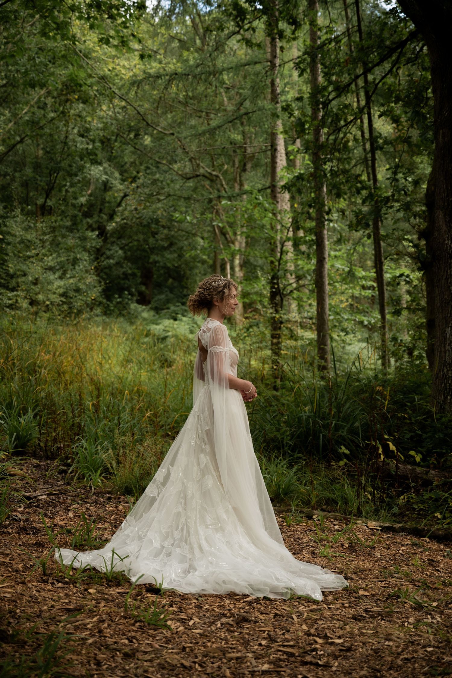 A bride in side profile looking out into the forest around her. Her hands are clasped at her waist and she wears a long white wedding gown.