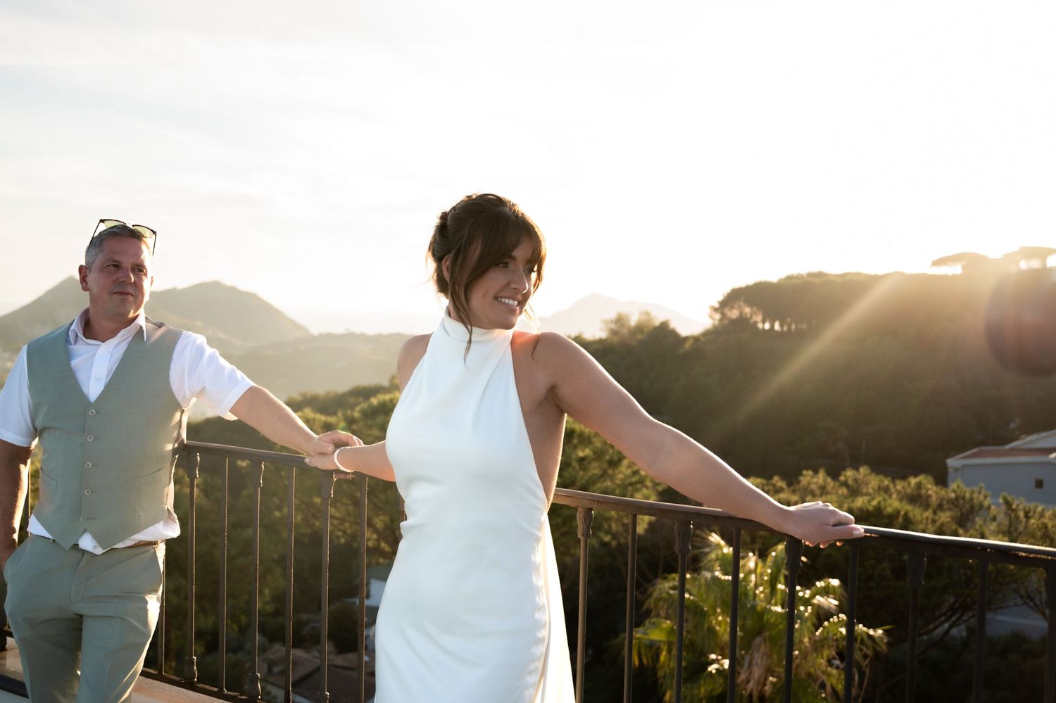 Couple hold hands and lean on railings of a rooftop terrace as they watch the sun setting in an orange hazy glow over the treetops below.