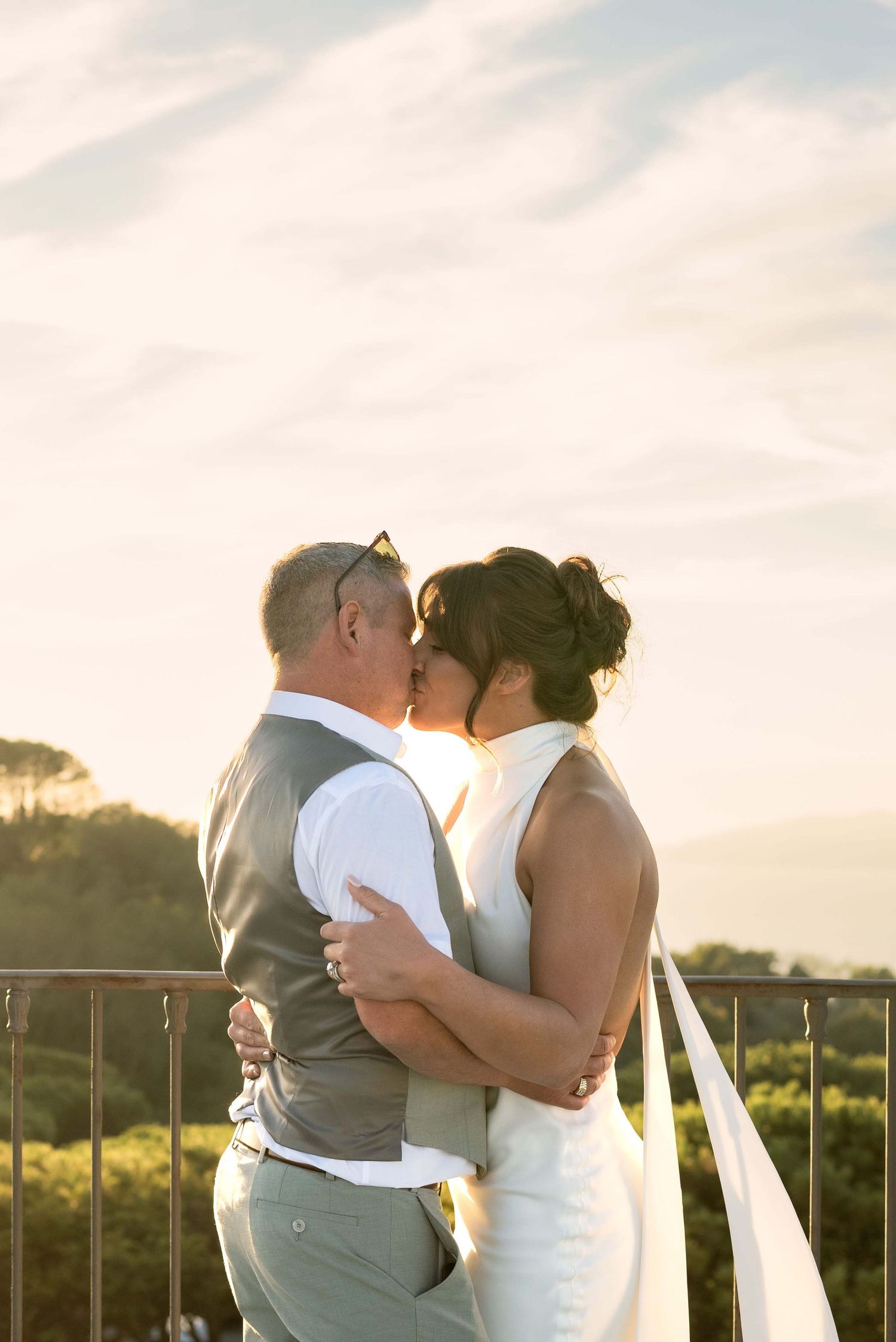 A bride and groom embracing and kissing on a balcony against a backdrop of sunlit trees at golden hour in Sorrento Italy.