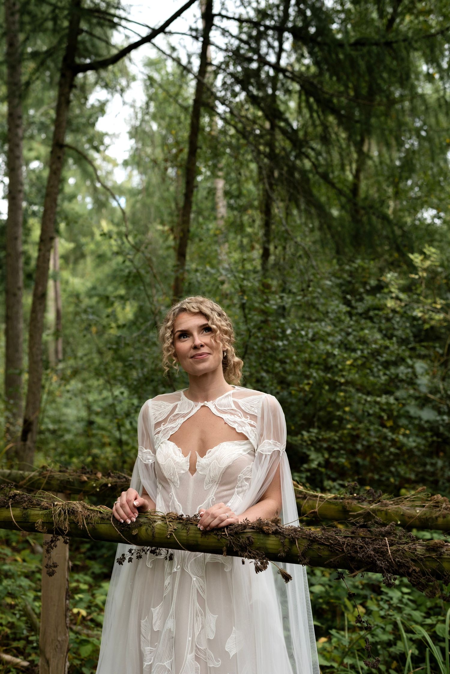 A woman in a caped corset gown leans on a bridge entwined with ivy in the forest. She looks up at the tall surrounding trees and smiles.
