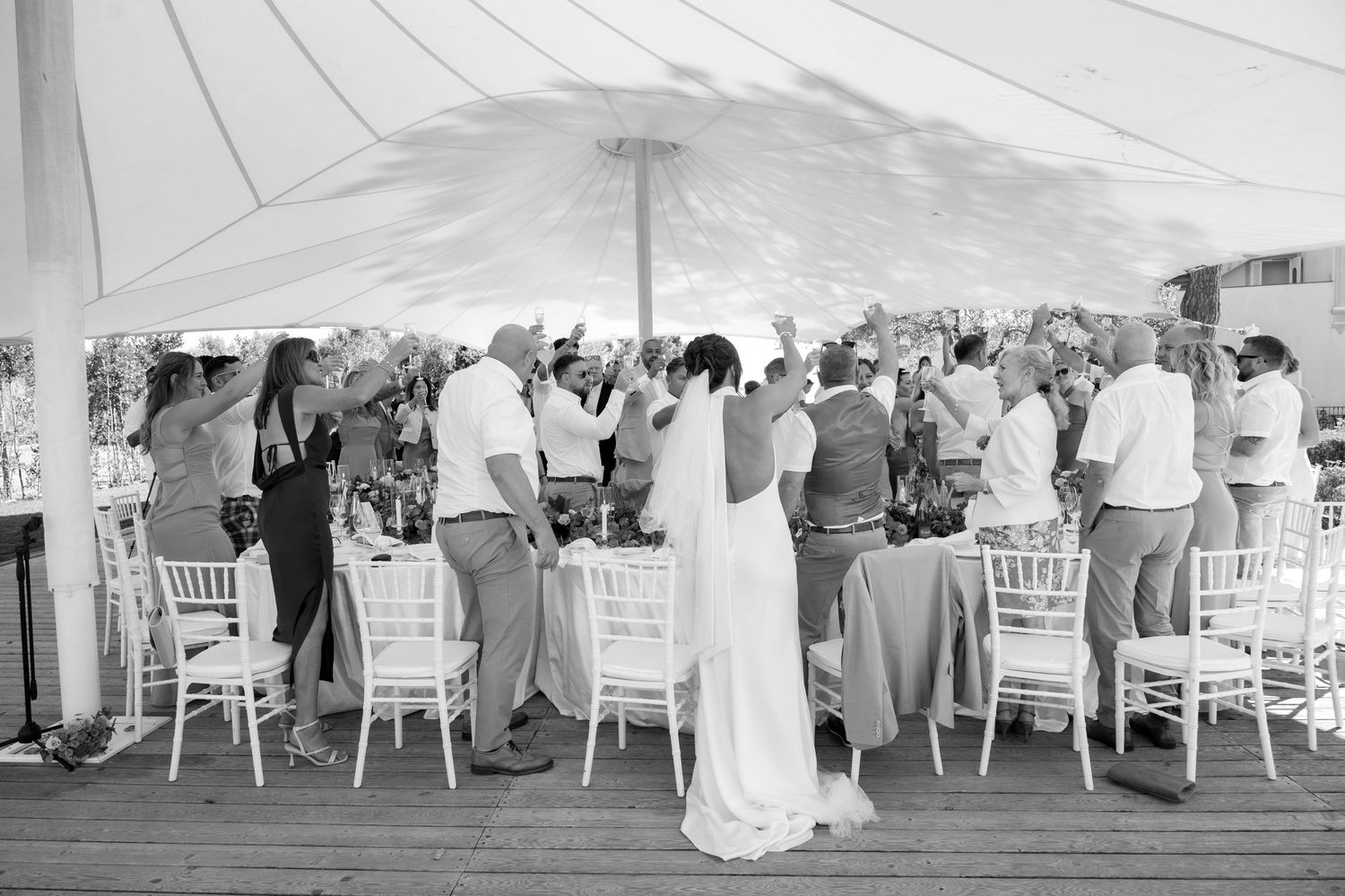 A party of happy wedding guests stand up from their seats and excitedly wave and clap to toast a speech.