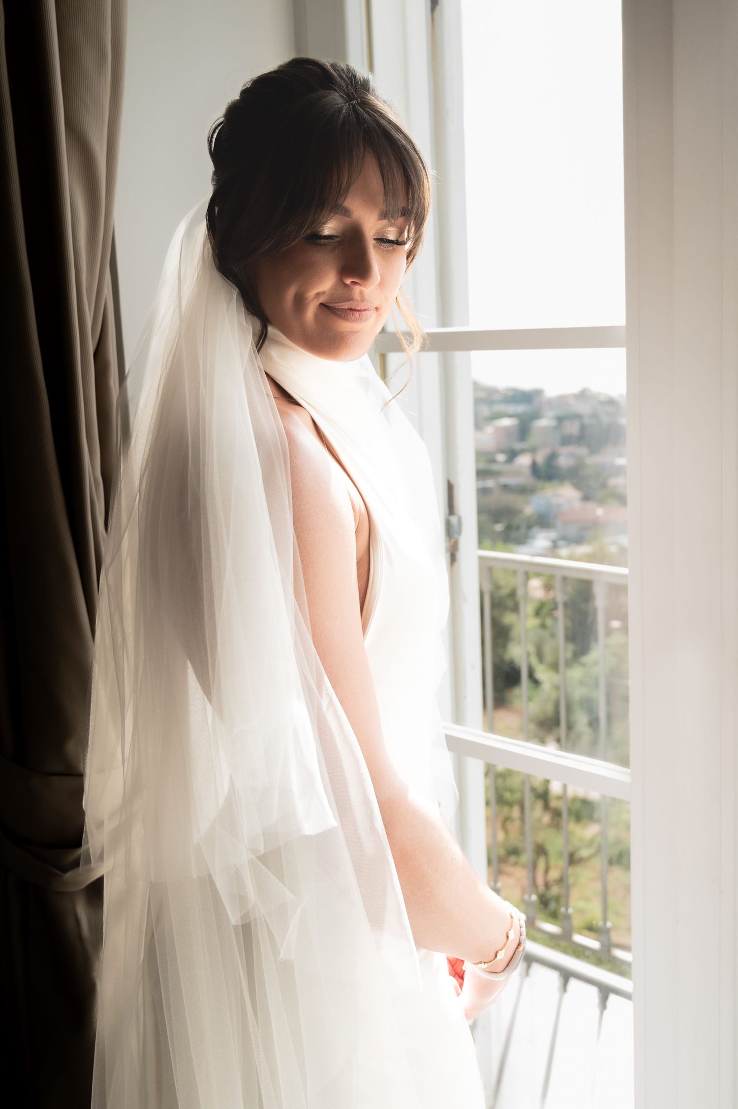 Beautiful woman stands in a window as the glow of sunshine casts over her face and silk gown. She has a relaxed smile and looks content.