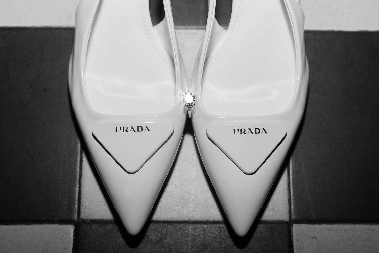A pair of white Prada slingback heels, photographed on a tiled floor. A sparkling diamond ring balances delicately between the shoes.
