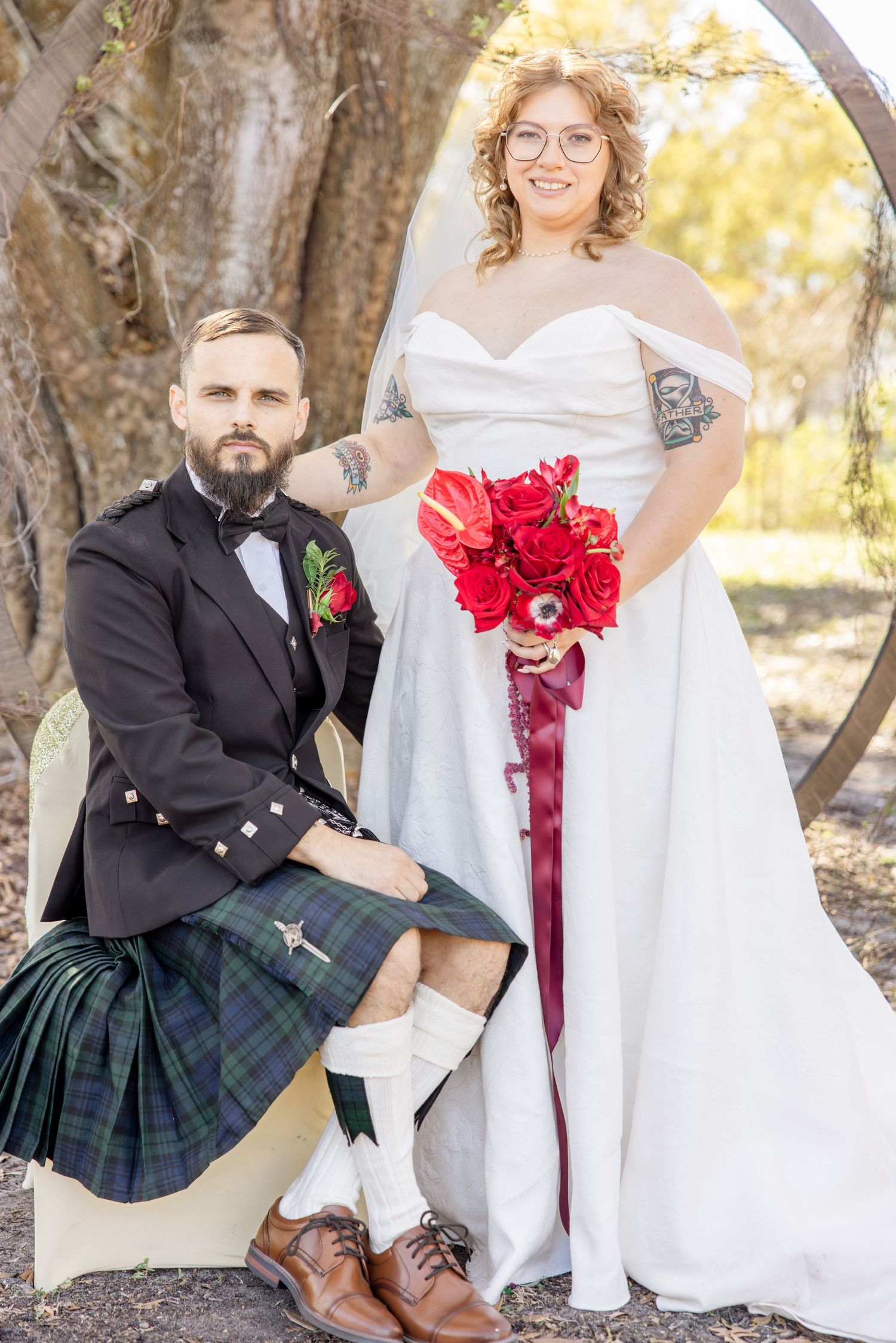 Groom in Scottish kilt and bride in white off-shoulder gown pose together outdoors with red rose bouquet.