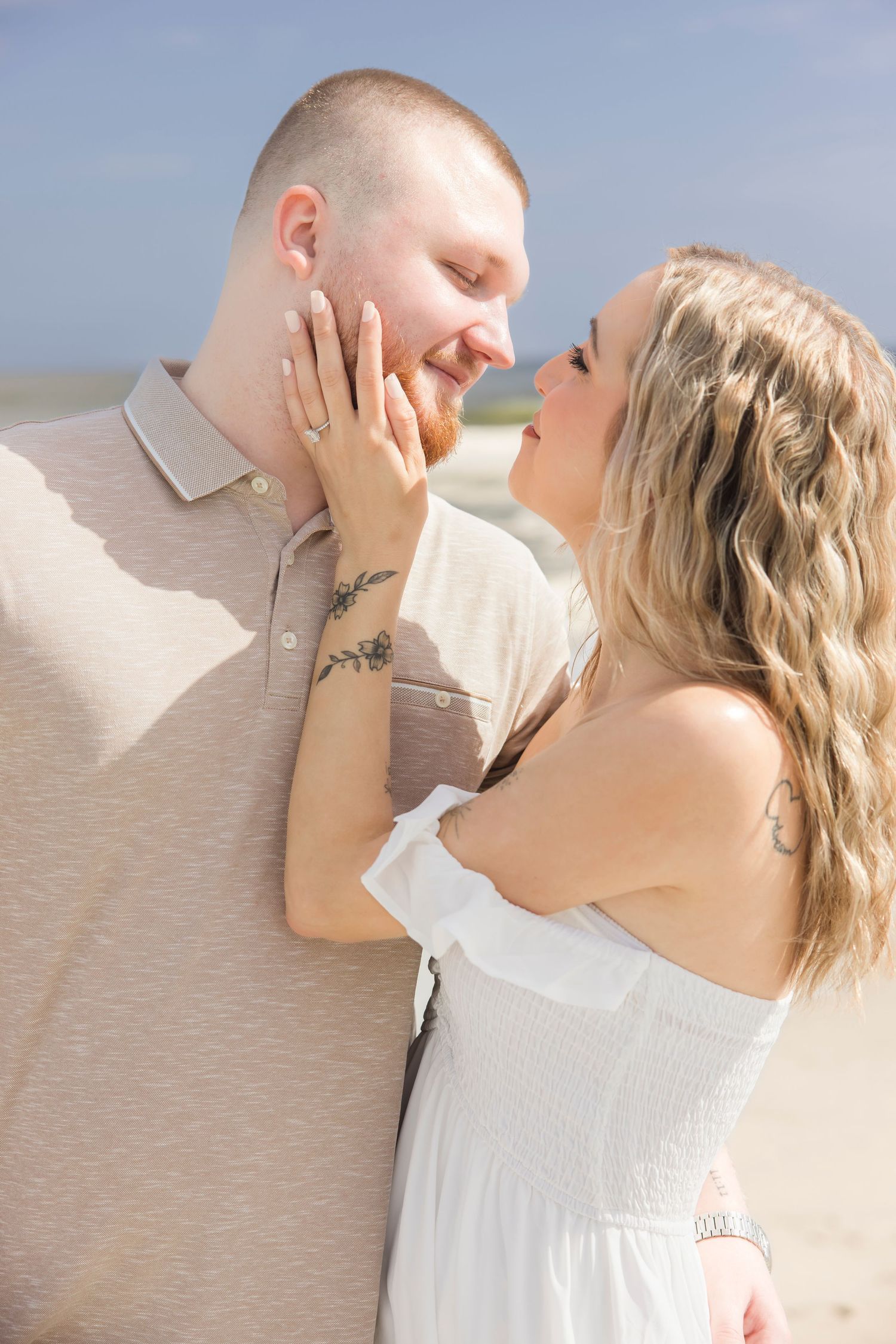 Couple sharing a romantic moment on the beach, woman in white dress touching man's face, both smiling.