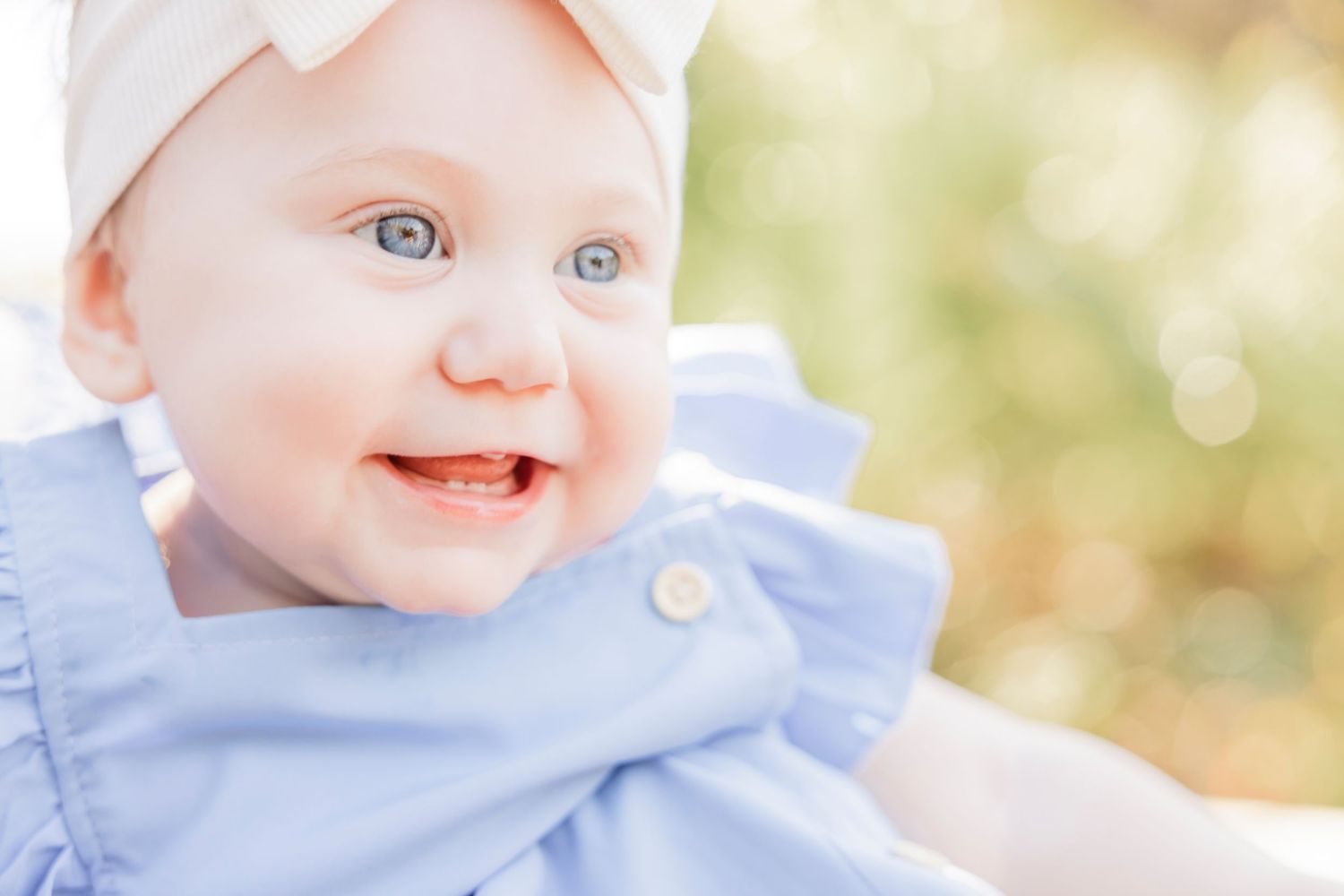 Smiling baby girl with blue eyes wearing a light blue dress and white bow headband outdoors.