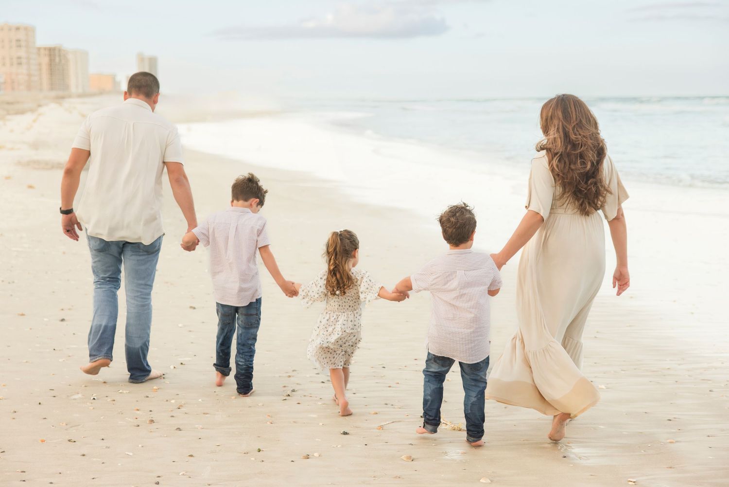 A family of five walks hand-in-hand along a sunny beach shoreline from behind.