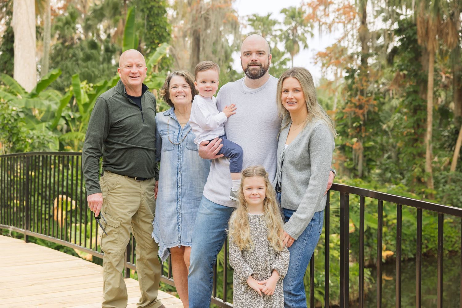 Multi-generational family of six smiling together on a wooden bridge surrounded by lush greenery.