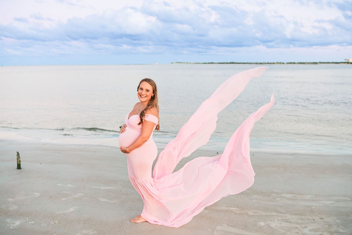 Pregnant woman in flowing pink off-shoulder gown standing in calm ocean water, fabric billowing.