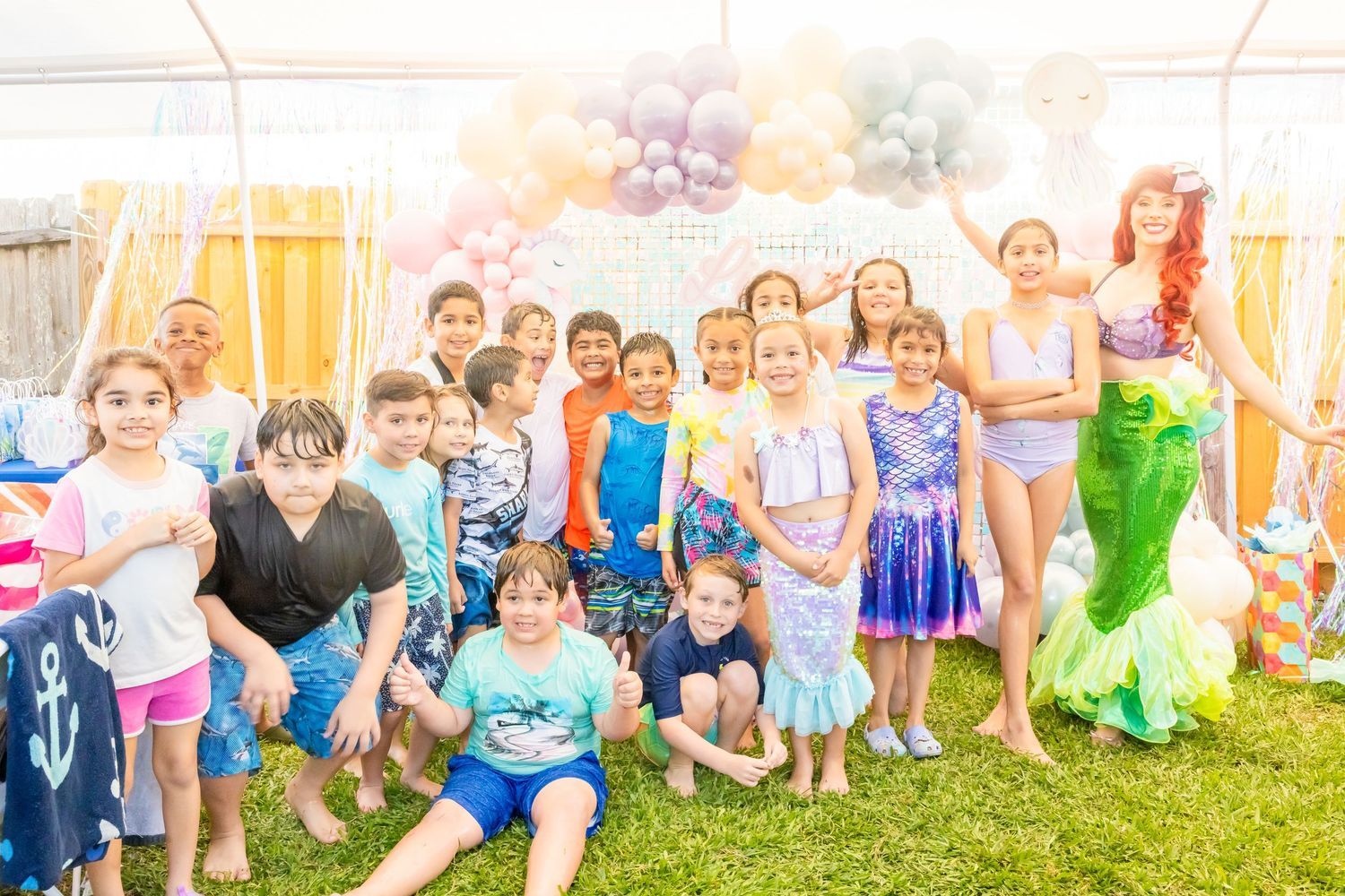 A large group of children pose outdoors with a Little Mermaid Ariel character at a birthday party with balloon decorations.
