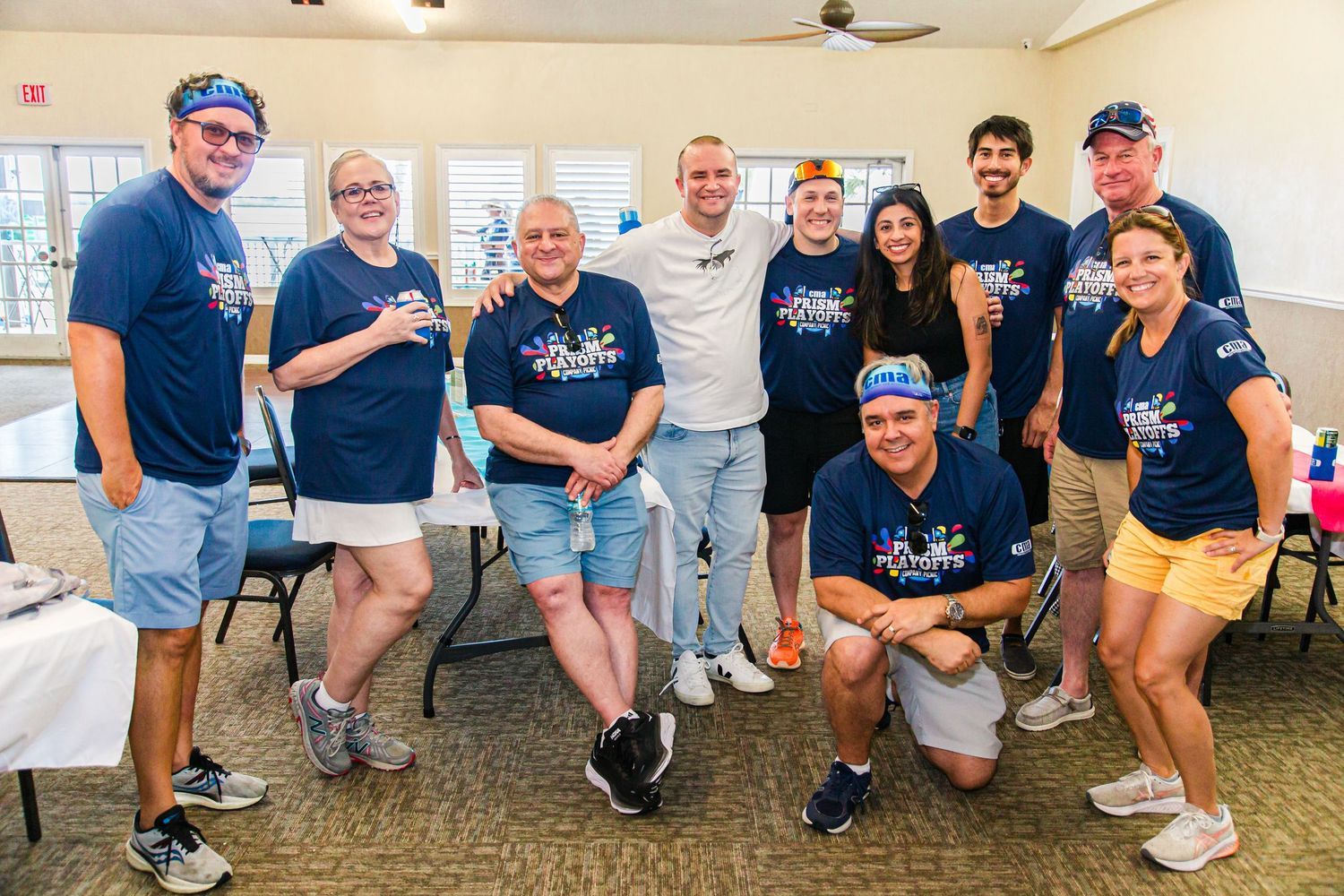 Group of nine people in Prism Playoffs t-shirts pose together smiling inside a community event hall.