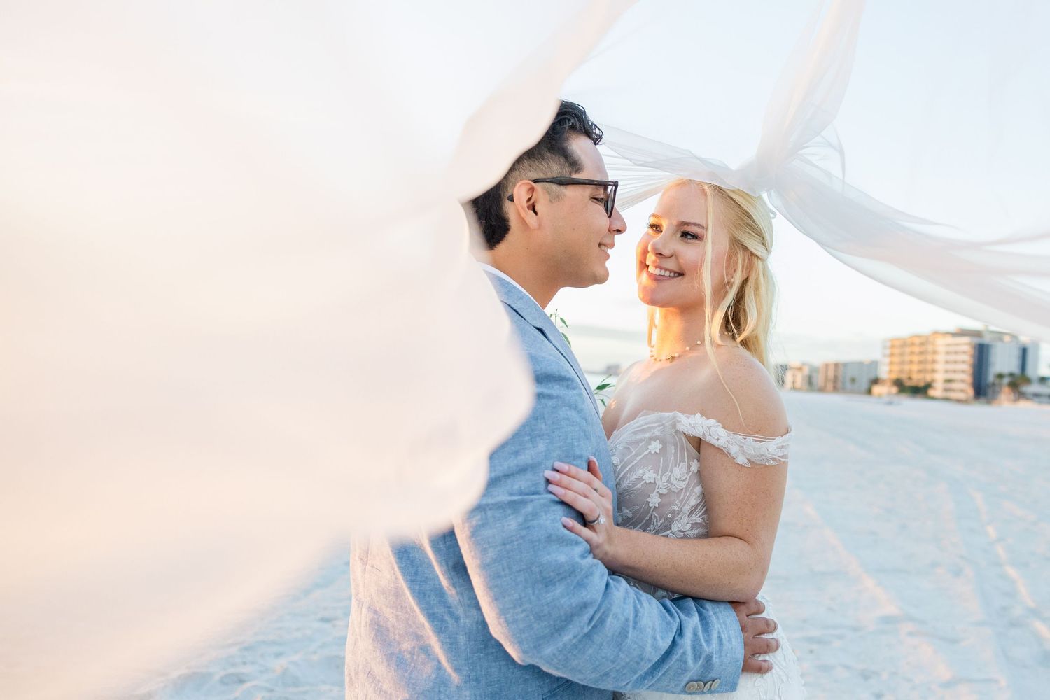 Couple embraces on beach with veil flowing in wind during romantic sunset wedding photo.