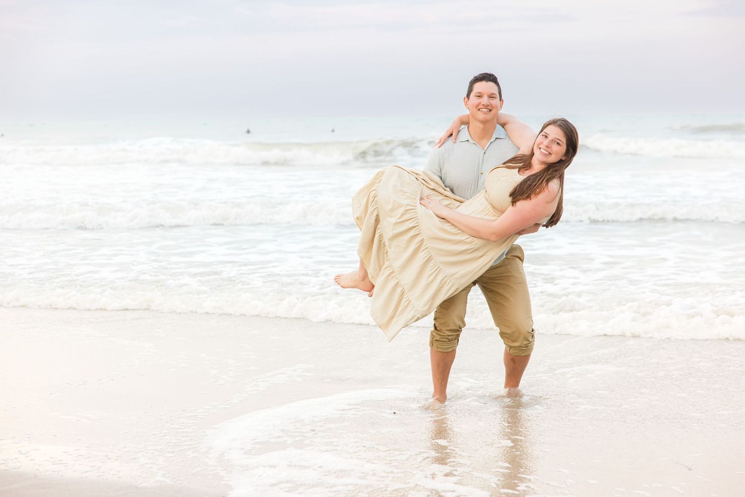 Couple laughing and embracing on a beach, man lifting woman in a flowing dress near ocean waves.