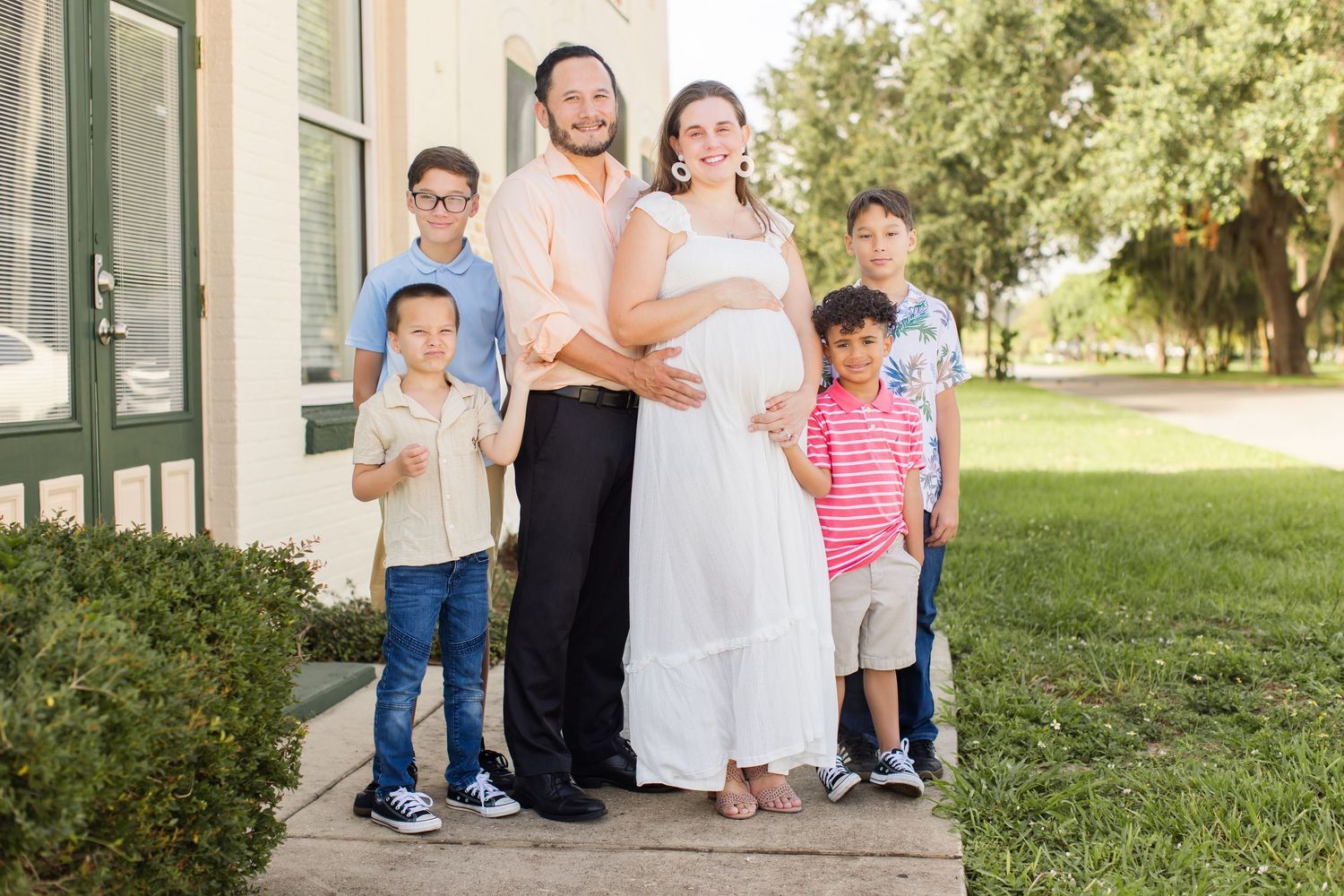 Pregnant woman in white dress with husband and three boys posing outside building on green lawn.
