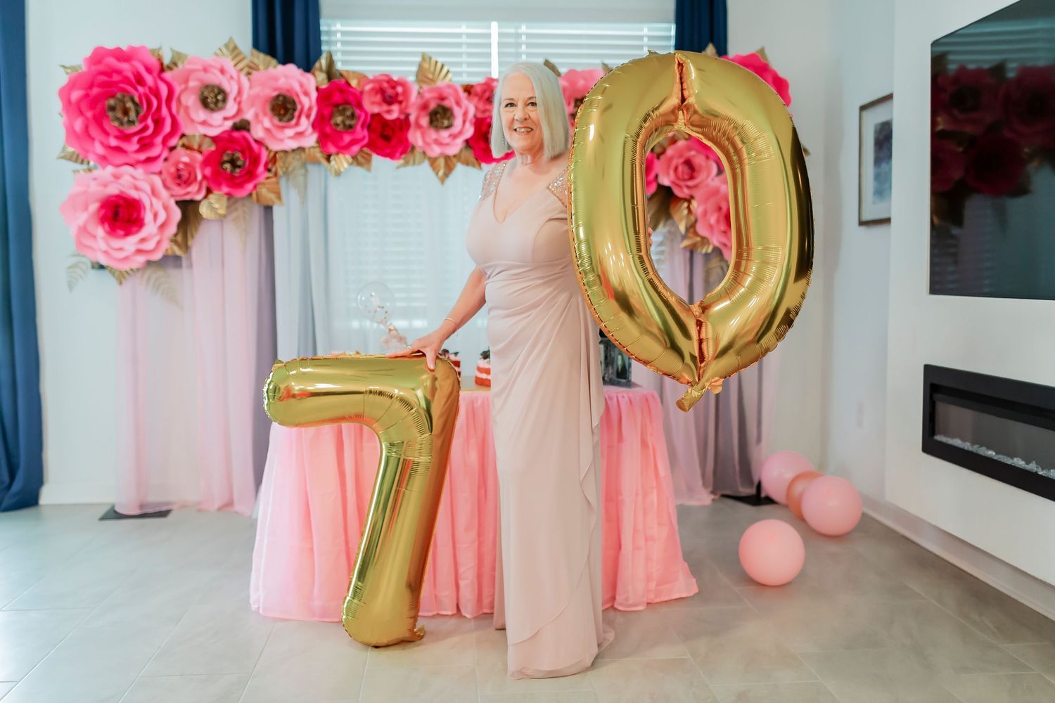 Woman celebrating 70th birthday holding large gold number balloons with pink floral backdrop.