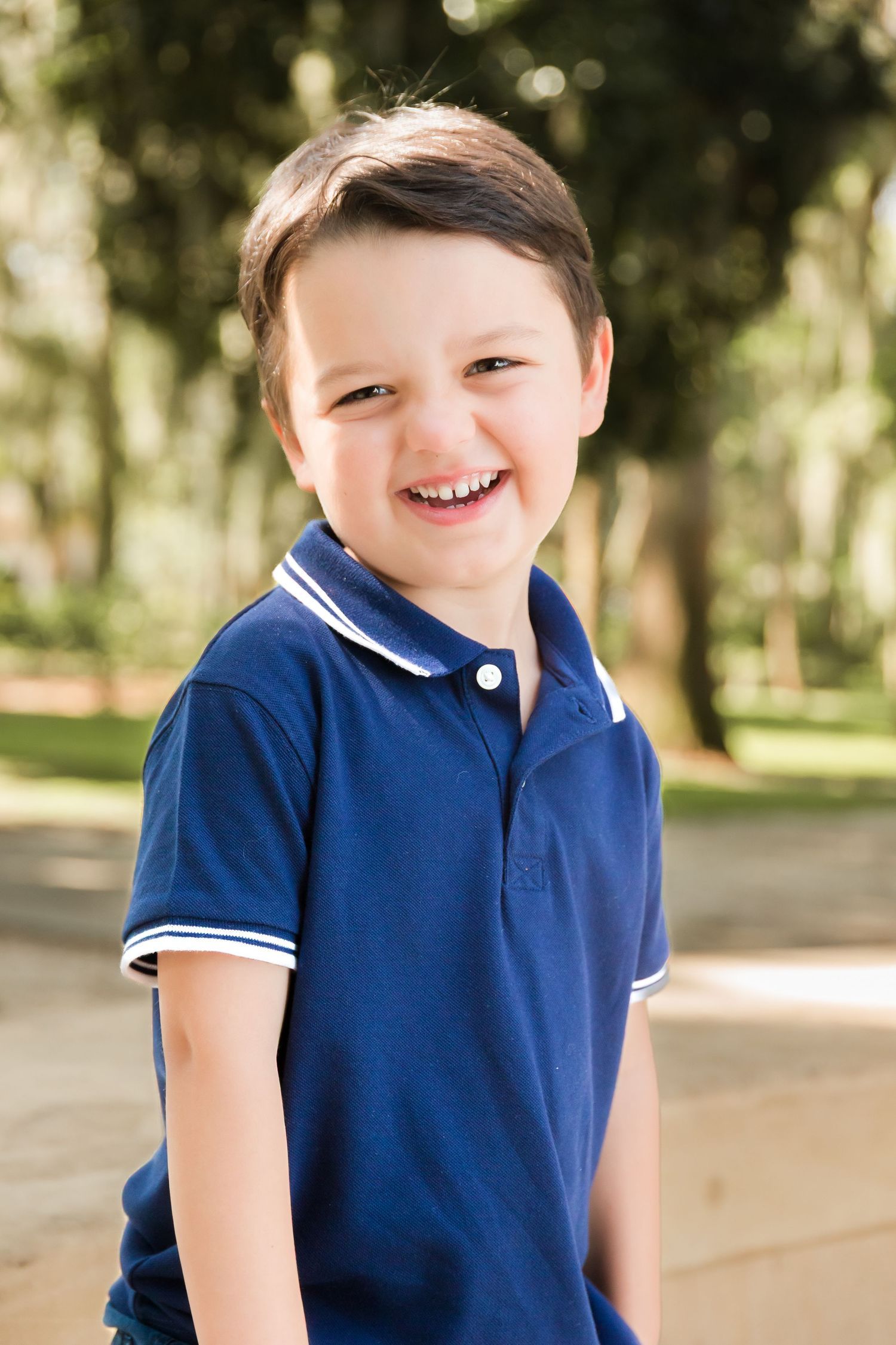 Smiling young boy in a navy blue polo shirt laughing outdoors in a sunny park setting.