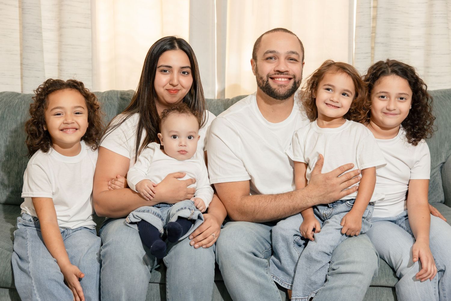 Smiling family of six in matching white shirts and jeans sitting together on a gray sofa at home.