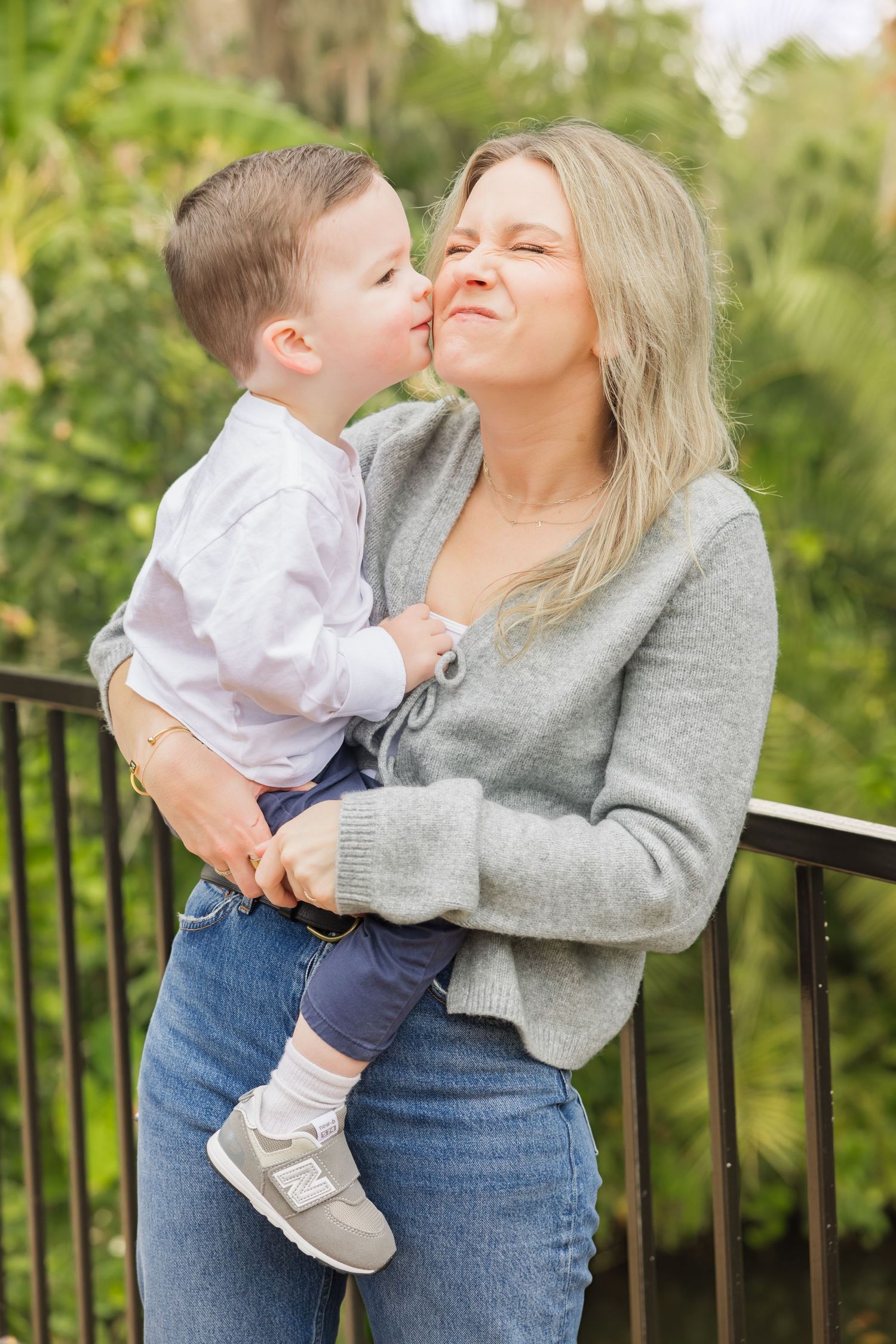 Blonde mother holding toddler son who gives her a sweet kiss on the cheek outdoors.