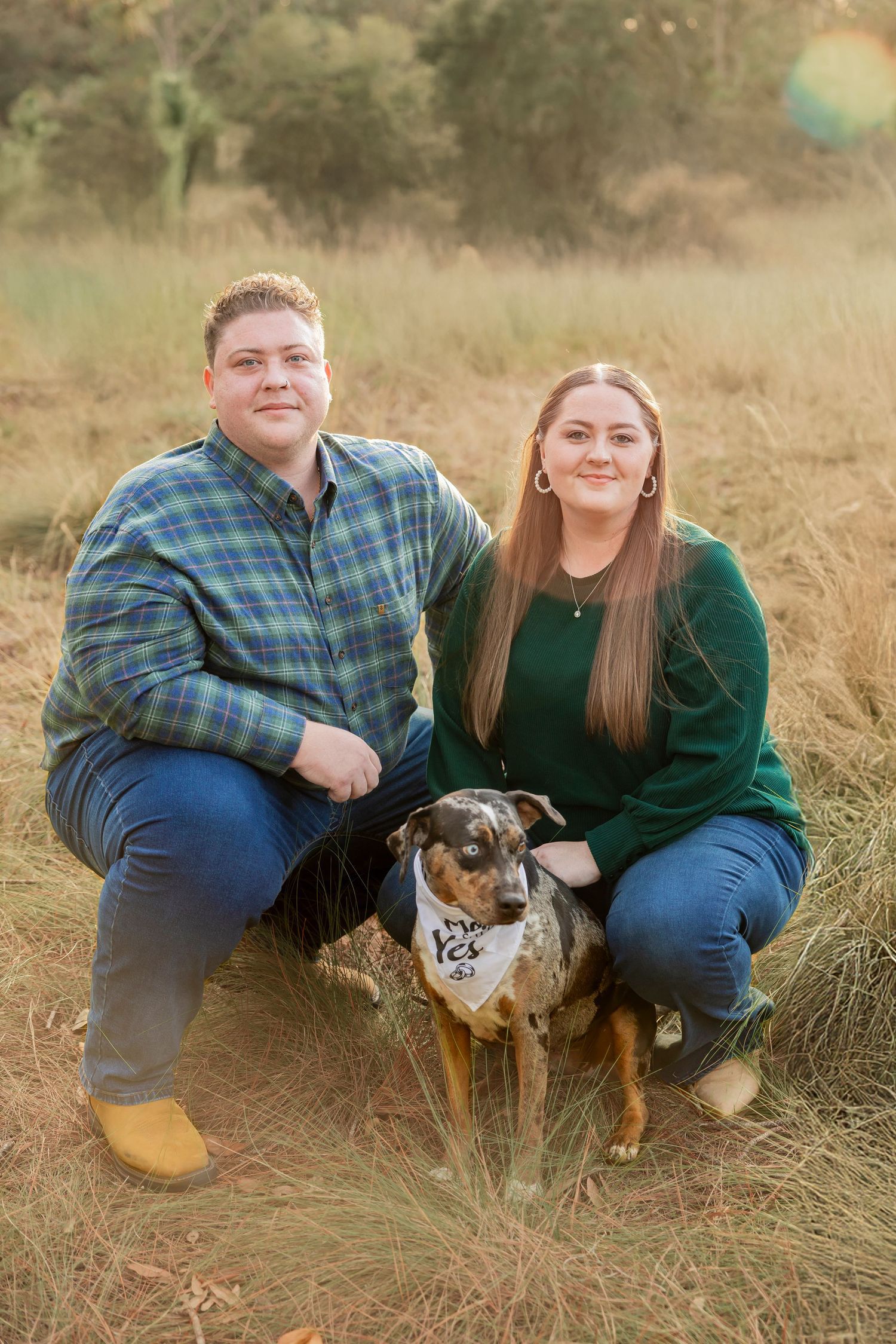 Smiling couple crouching with their dog wearing a bandana in a golden field at sunset.