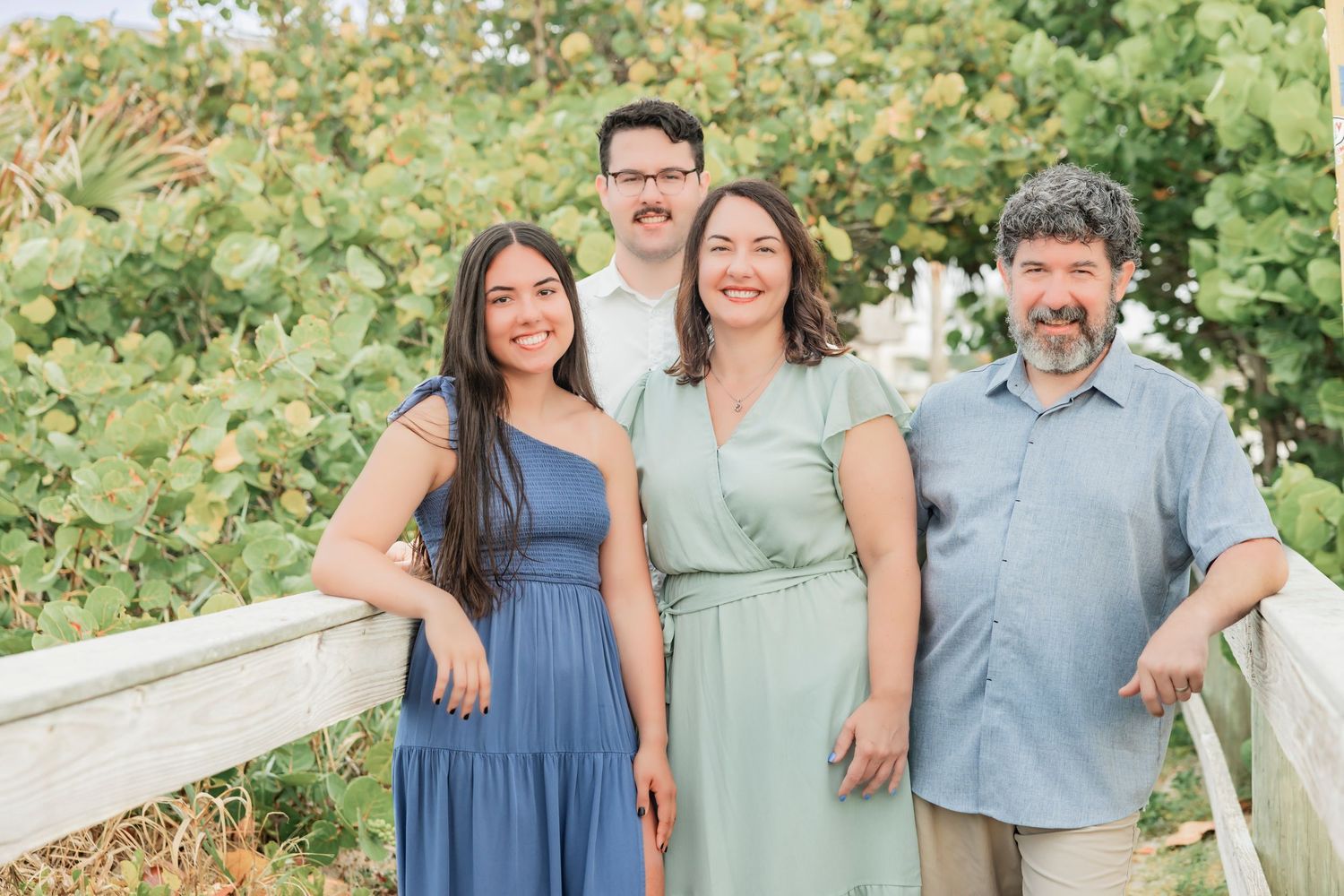 Family of four smiling outdoors near a white fence with lush green trees in the background.