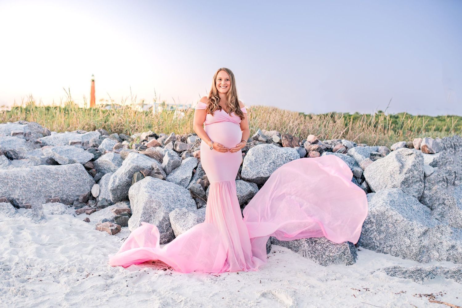 Pregnant woman in flowing pink gown poses on sandy beach near rocks.