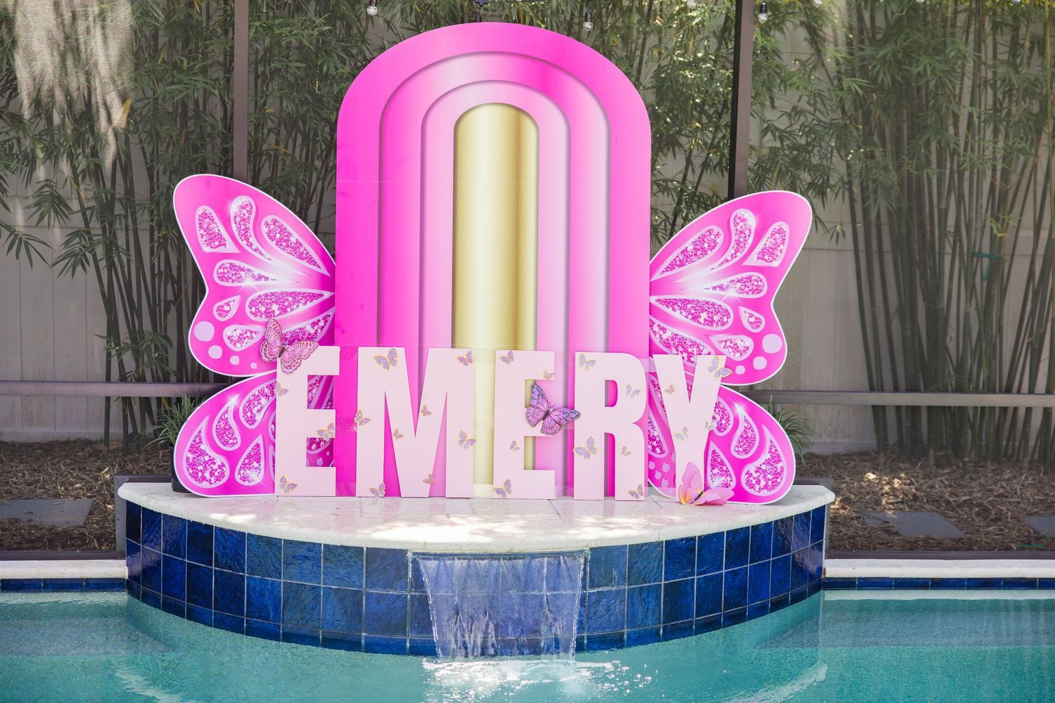 Pink butterfly-themed EMERY name sign with gold arch displayed on a pool fountain for a birthday party.