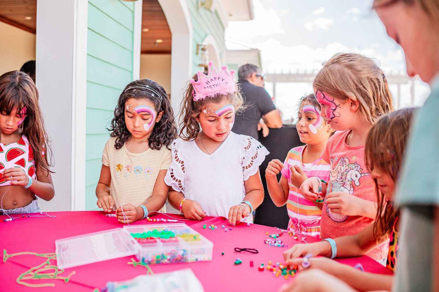 Girls with face paint make jewelry at a pink table during a birthday party craft activity.