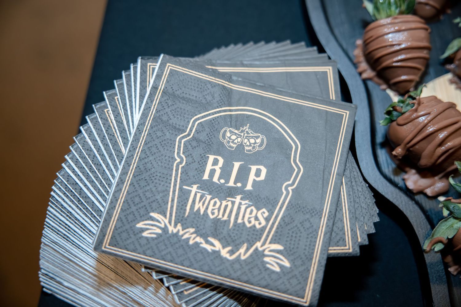 Stack of RIP Twenties tombstone themed gray cocktail napkins beside chocolate covered strawberries at party.