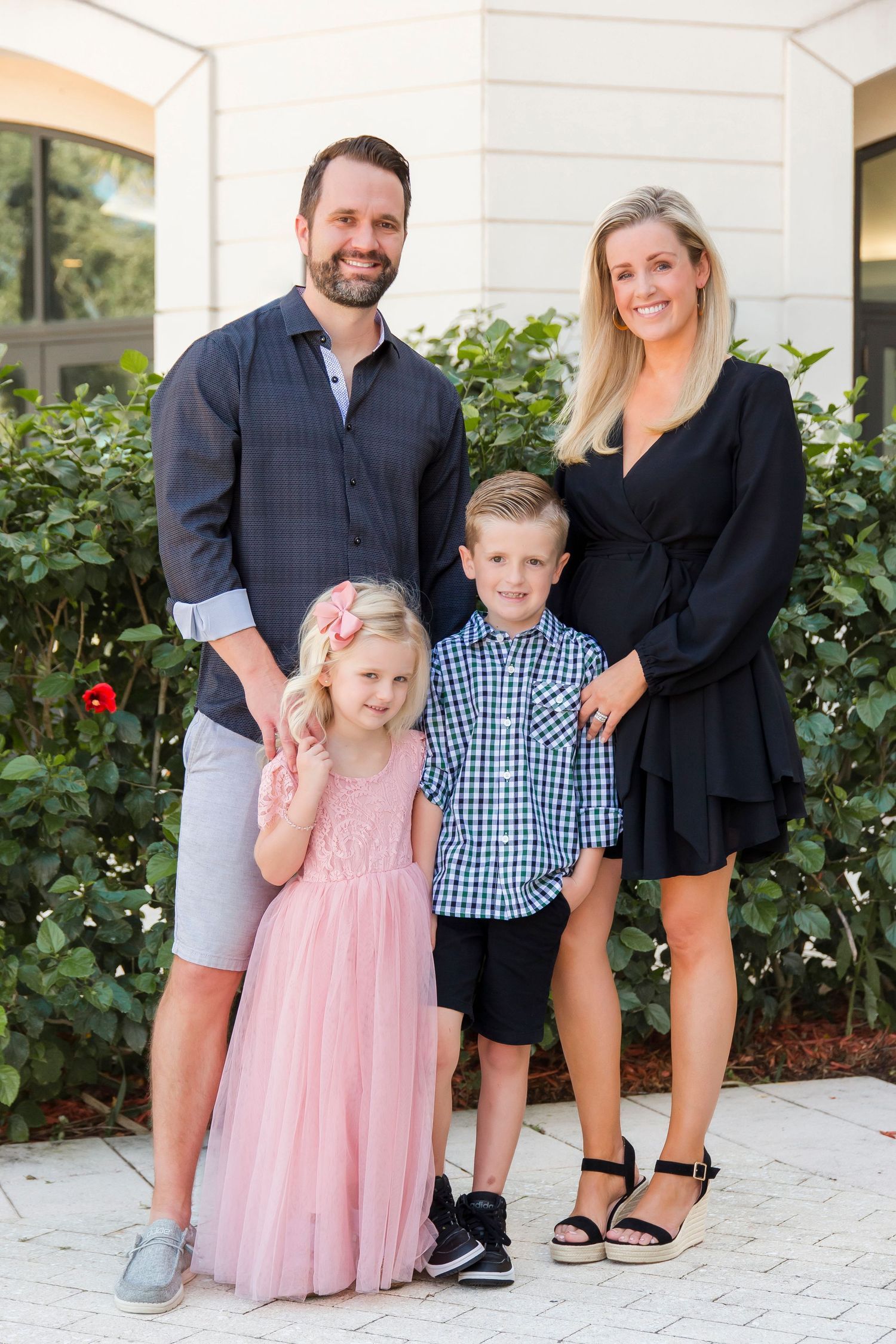 Family of four posing outdoors, parents in dark outfits with daughter in pink tutu dress and son in plaid.