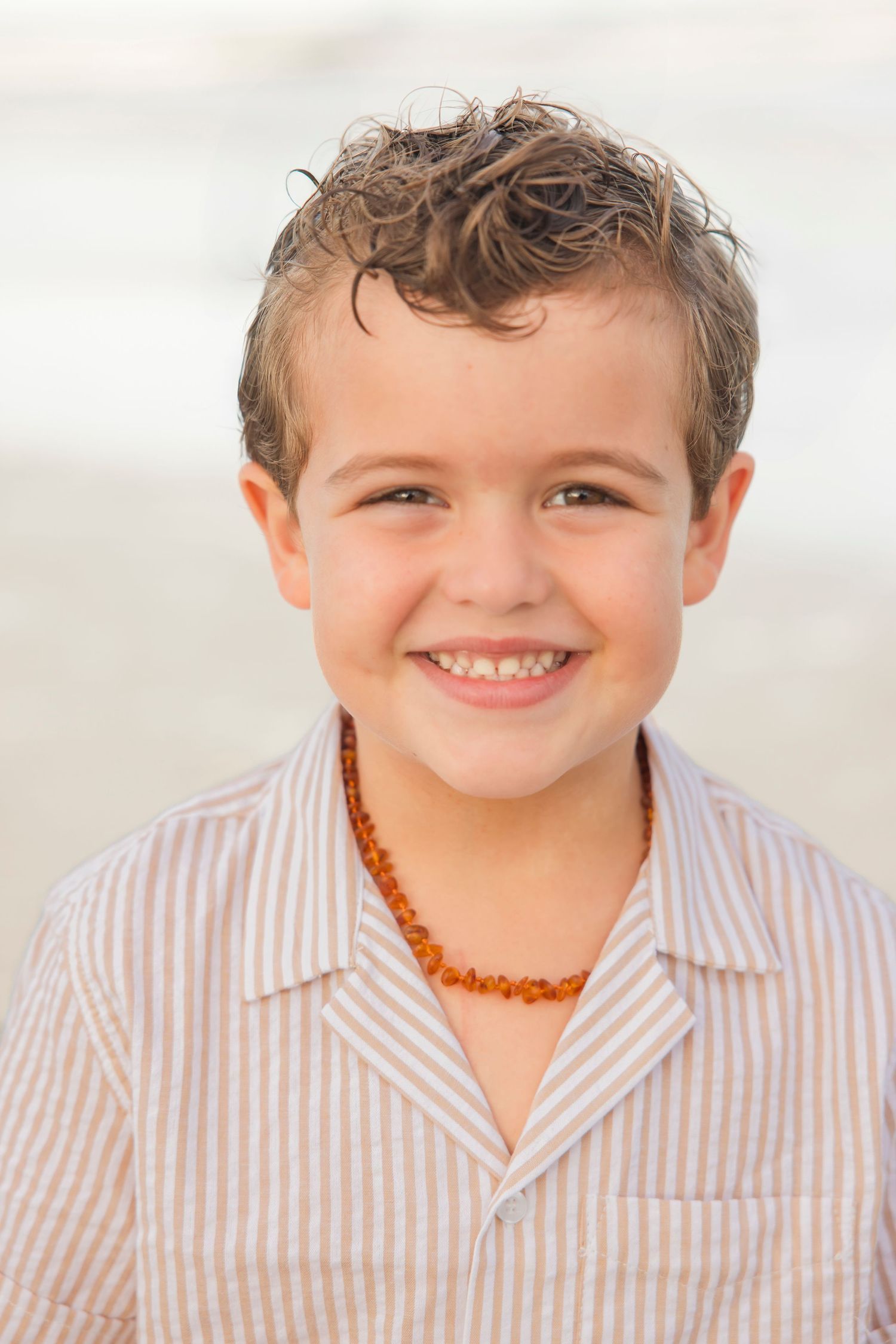 Adorable young boy with curly hair and amber bead necklace smiling brightly at beach.