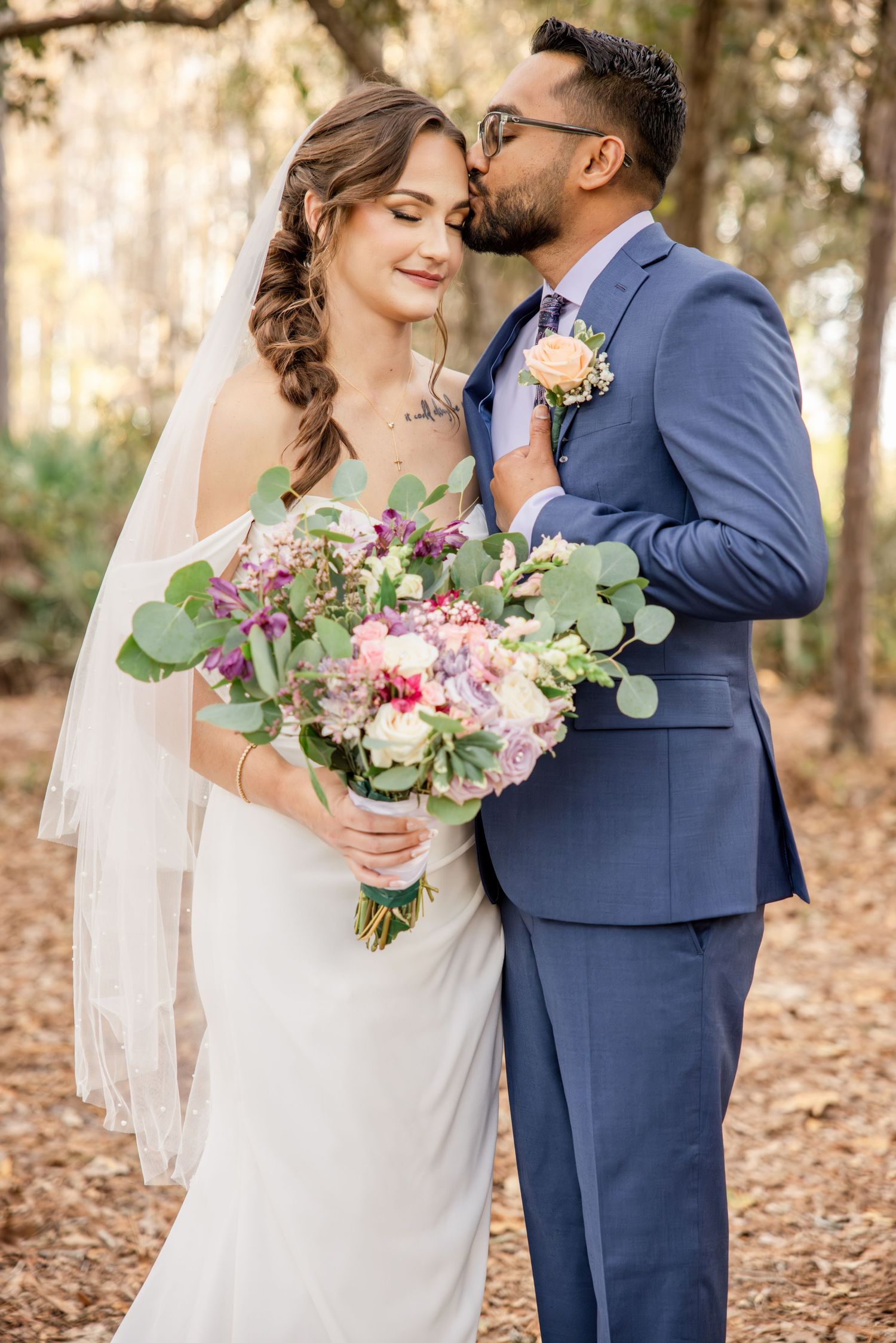 Groom in blue suit kisses bride's cheek as she smiles holding colorful floral bouquet in wooded outdoor setting.