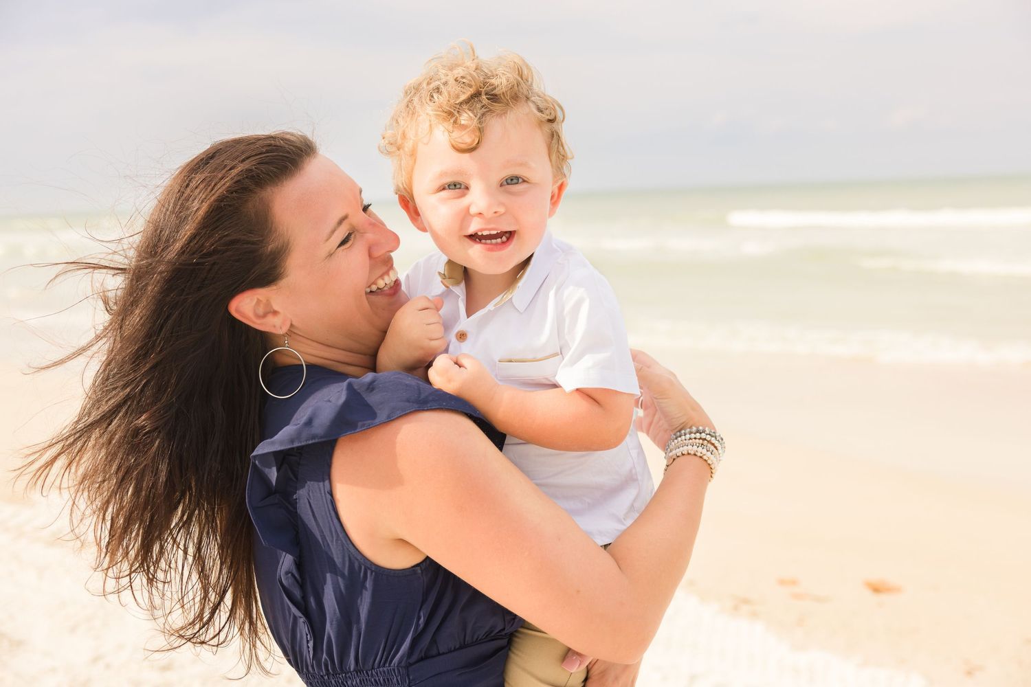 A smiling mother holds her curly-haired toddler on a breezy beach with ocean waves behind.
