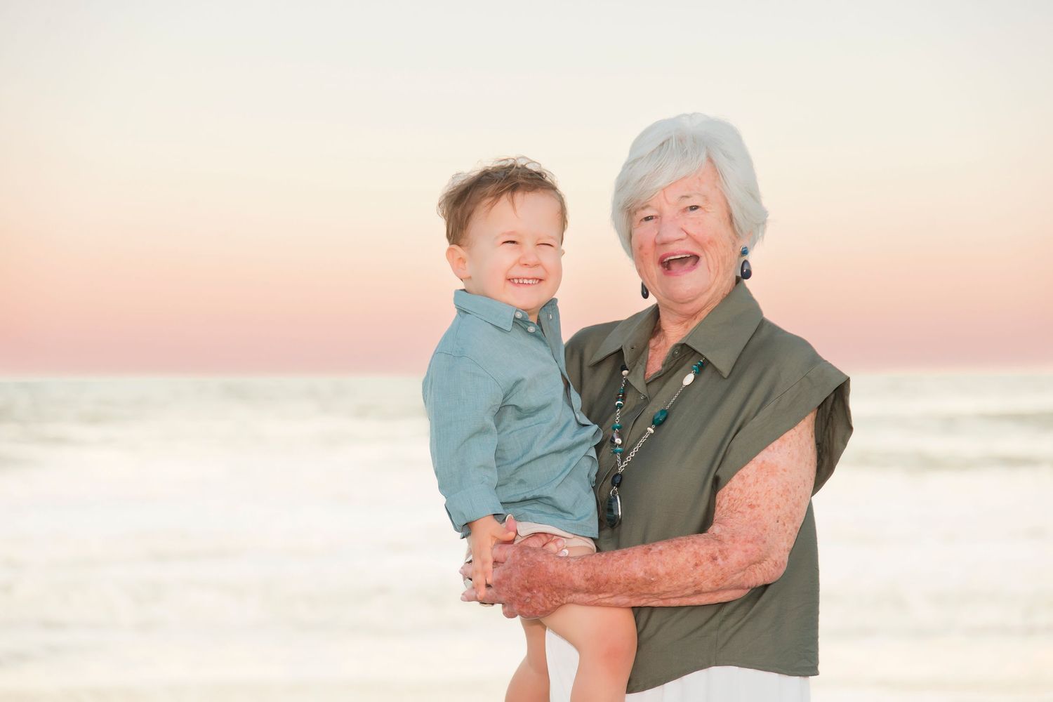 Grandmother laughing while holding a smiling toddler boy on a beach at sunset.