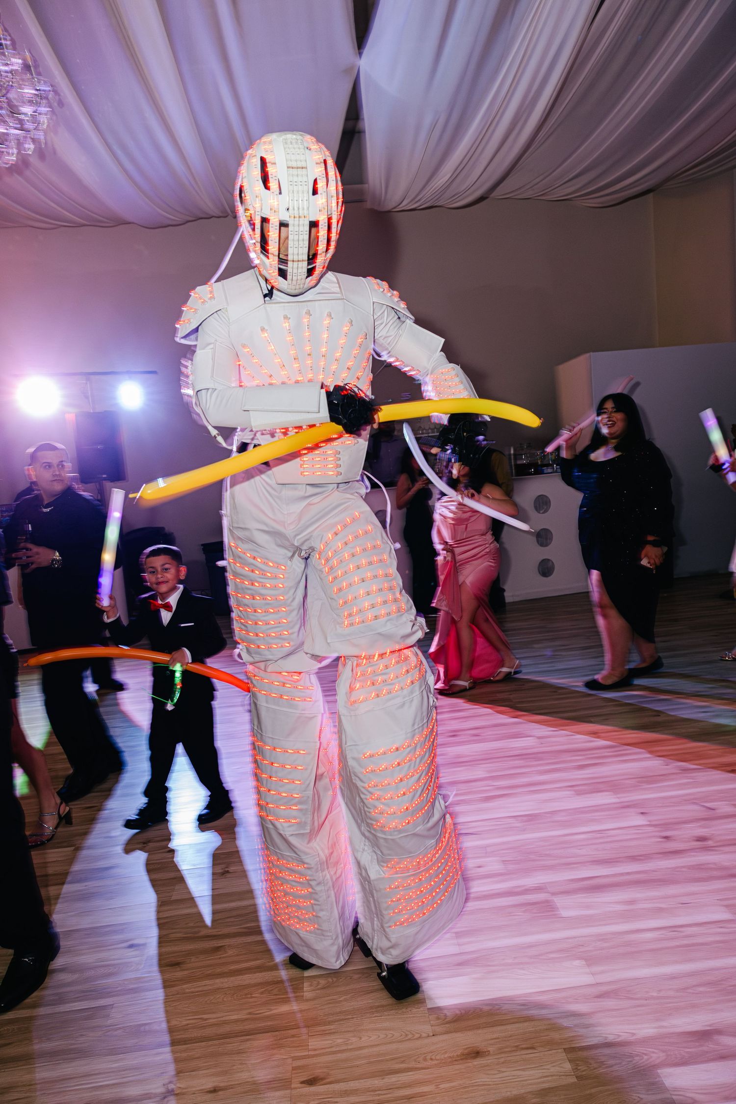 Performer in illuminated LED robot costume on stilts with hula hoops entertains guests on dance floor.