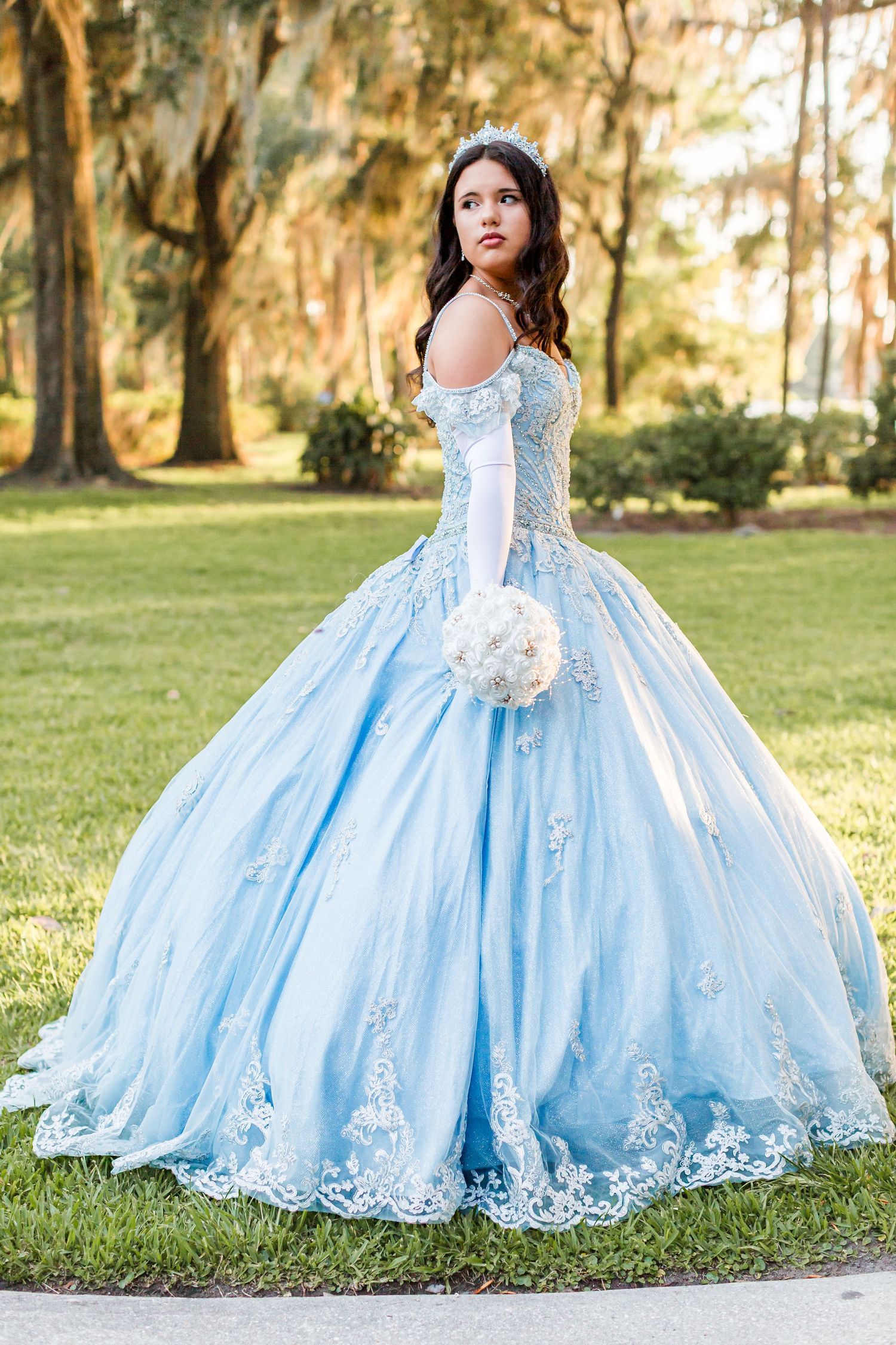 A young woman in a light blue quinceañera ball gown holds white flowers in a scenic park.