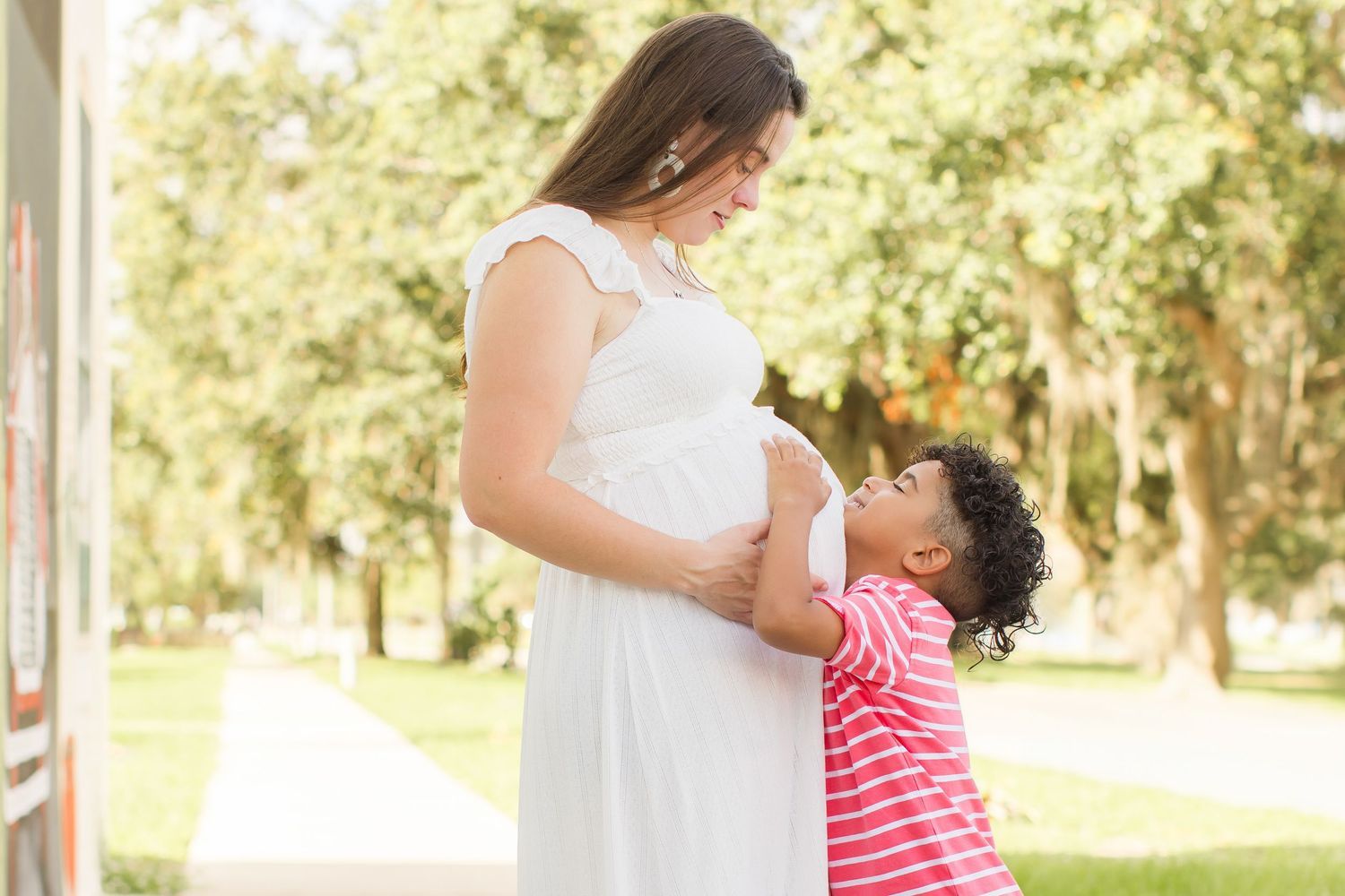 Young child hugging and kissing pregnant mother's belly outdoors in sunny park setting.
