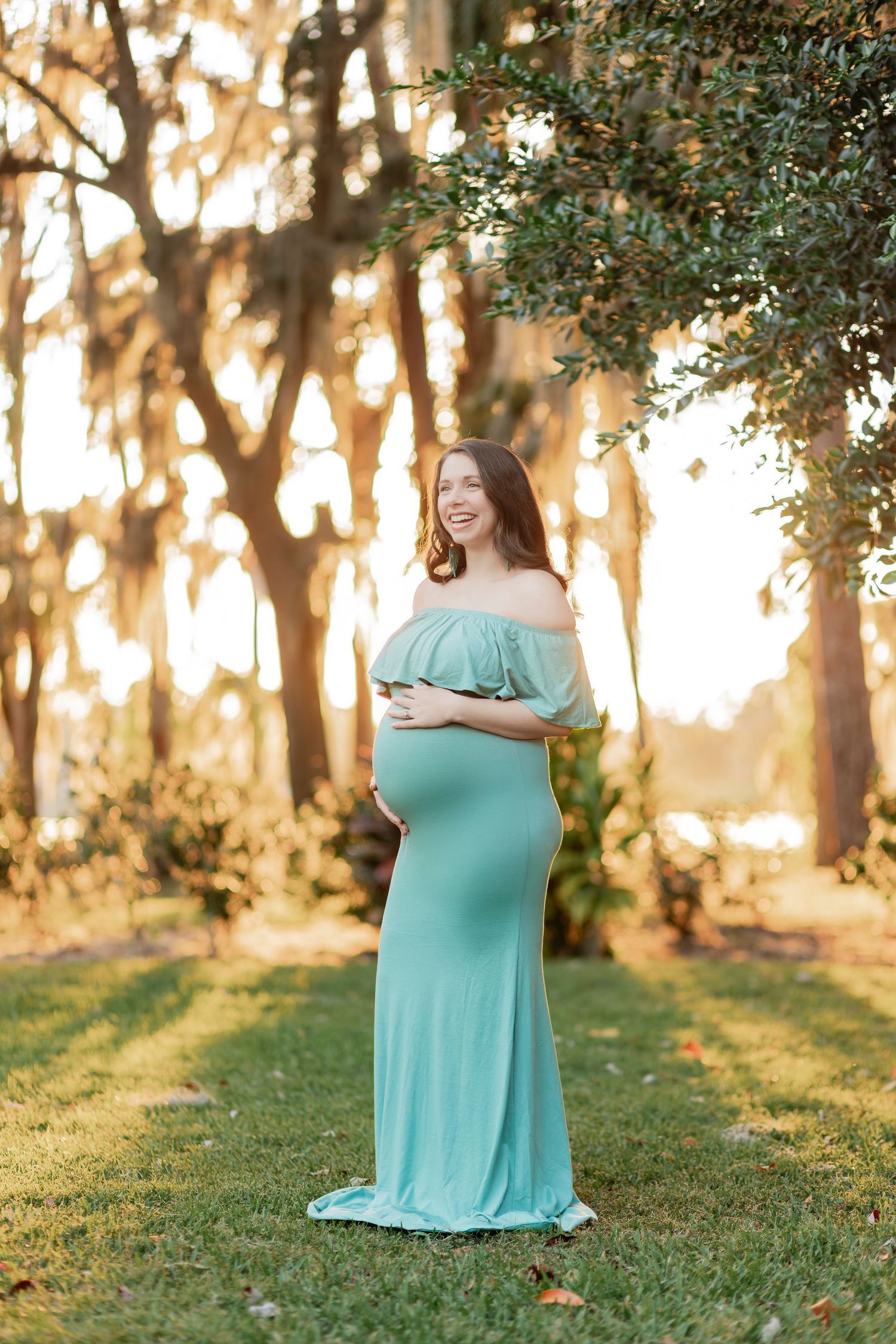 Smiling pregnant woman in teal off-shoulder maternity gown standing outdoors among trees at golden hour.