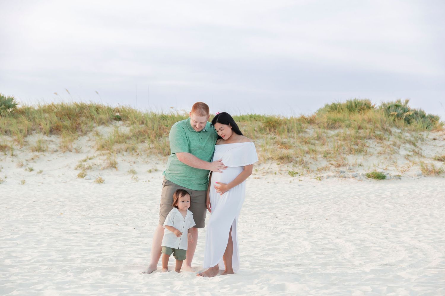 Family maternity photo with couple and toddler on white sandy beach.