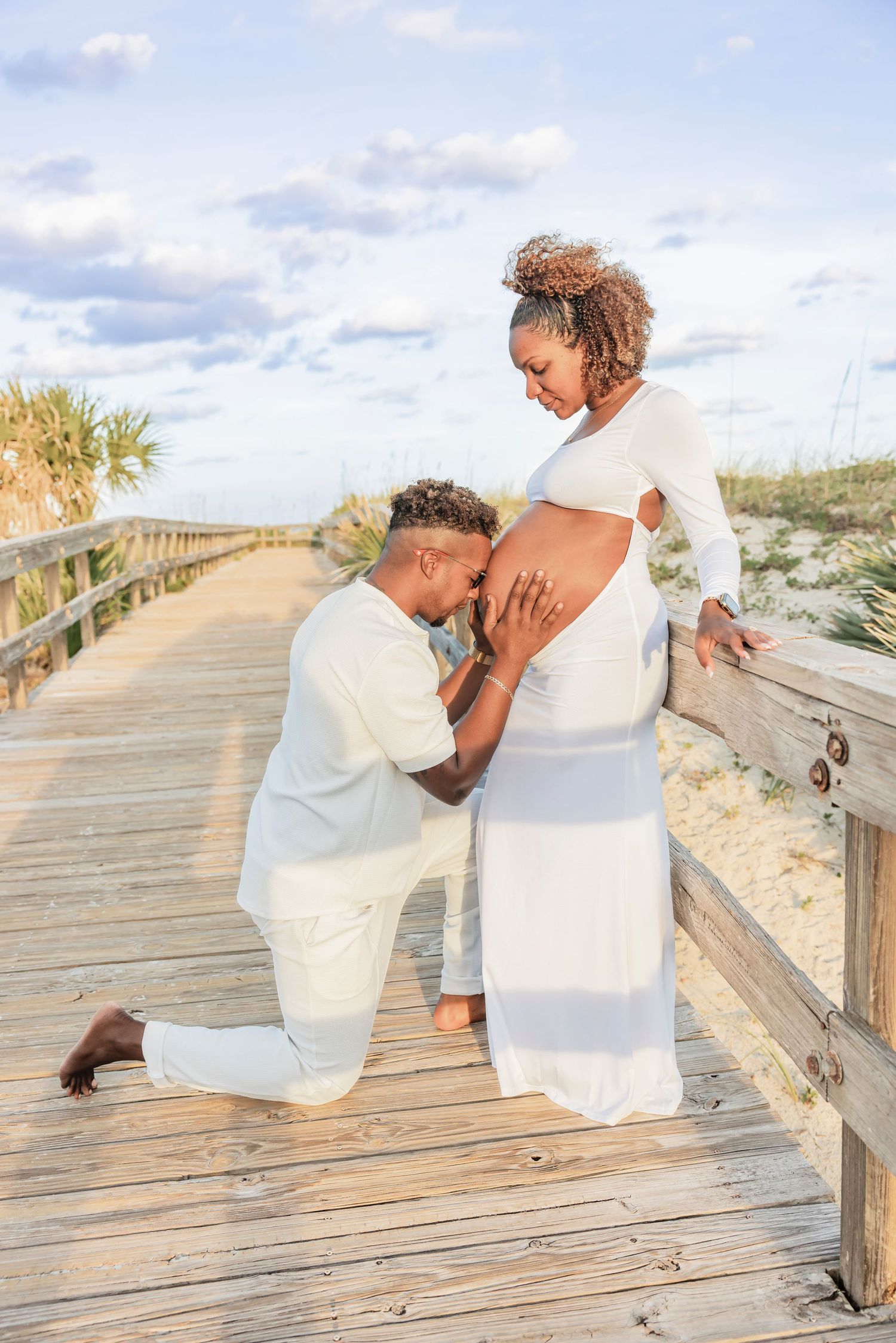 Man kneeling on a boardwalk kissing pregnant partners baby bump during a golden hour maternity photo session.