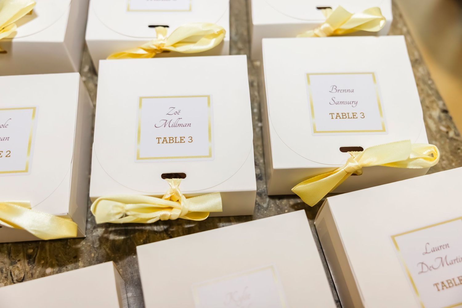 White gift boxes with gold labels and yellow ribbon bows arranged as wedding table seating cards on a surface.