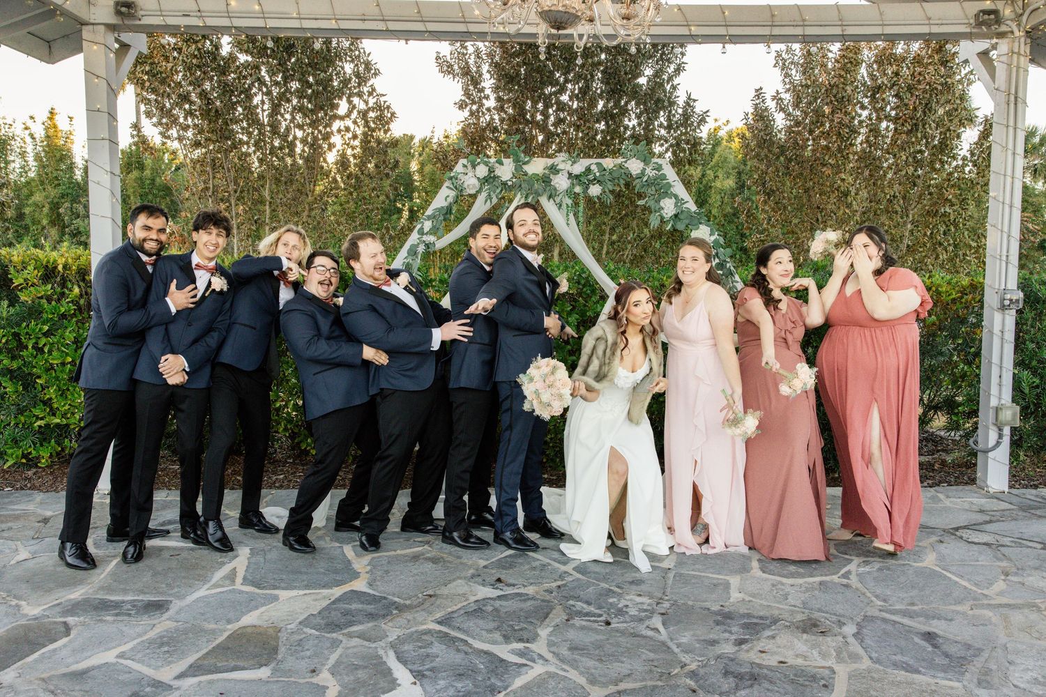 Wedding party poses joyfully under floral arch, groomsmen in navy suits and bridesmaids in blush and mauve gowns.