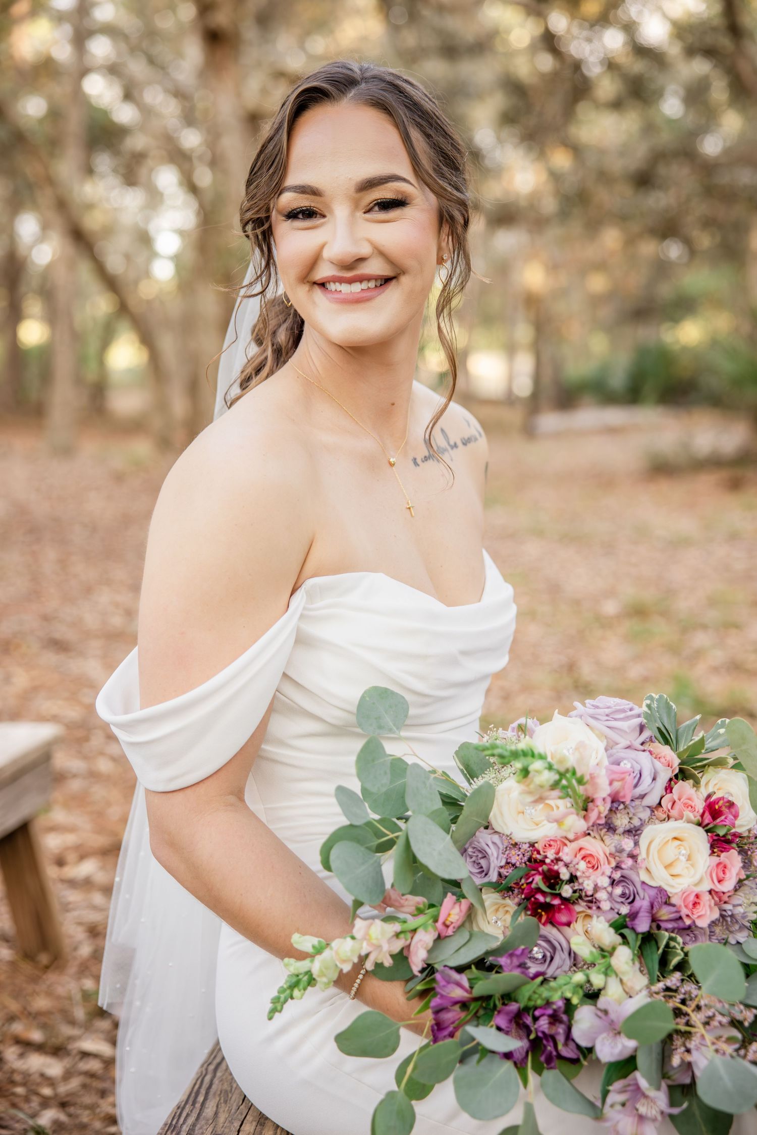 Smiling bride in off-shoulder white gown holding a lush purple and pink bouquet in a sunlit forest.
