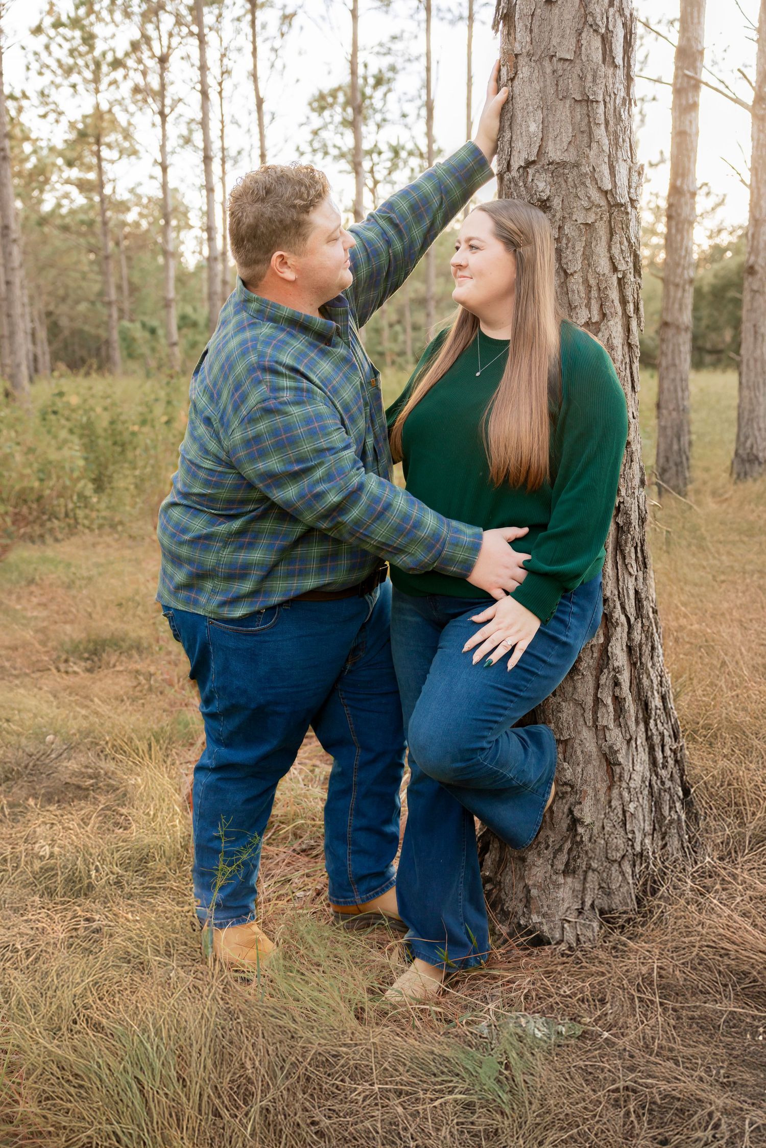 Couple sharing a romantic moment leaning against a pine tree in a wooded outdoor setting.